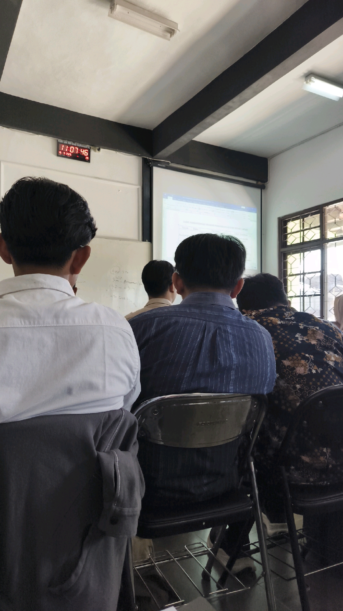 The image captures an indoor scene, likely a classroom or lecture hall, in Kartasura, Indonesia. The perspective is from behind several individuals seated, facing a projector screen. In the foreground, a person wearing a white collared shirt is visible from the back. To their right, another person in a blue striped collared shirt is seated. Further to the right, a person with a dark patterned shirt is also seated. The room features a projector screen displaying some text, whiteboards on the wall, and windows with visible gratings. A digital clock on the wall displays "11:07:46". The lighting appears to be from overhead fluorescent fixtures. The overall environment suggests an educational or presentation setting.