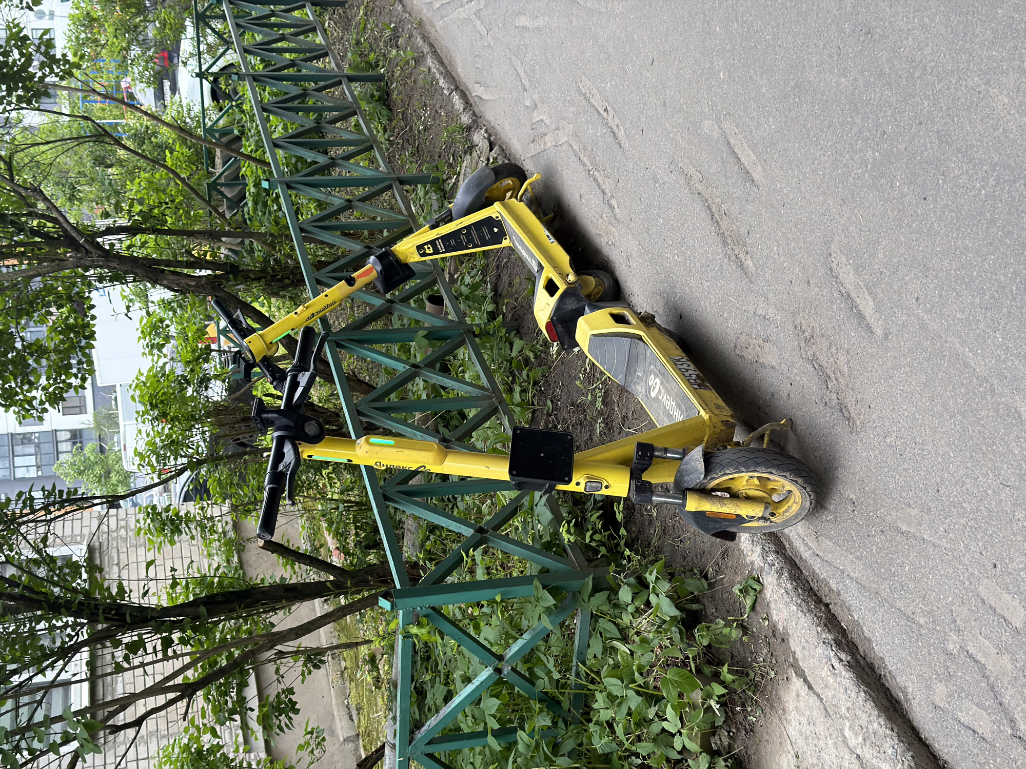 The image displays two bright yellow electric scooters positioned outdoors next to a green metal fence. Lush green trees and shrubs are visible behind and around the fence, with residential or urban buildings partially discernible through the foliage in the background. An asphalt path or road runs parallel to the fence. One scooter leans against the fence, while the other lies on its side, partially resting on the ground and against the fence, suggesting they are parked or discarded.
The scene is set during the daytime, under what appears to be an overcast or cloudy sky, indicated by the diffused lighting and lack of strong shadows. The scooters show signs of regular use, with some dirt on their wheels and lower parts. The asphalt path features some irregular indentations.
Prominently displayed on the side of both scooters is the brand name "Яндекс Go" (Yandex Go), identifying them as part of a shared electric scooter service. A partial alphanumeric code, "B53K", is discernible on the rear of one scooter. No people are present in the image. The setting appears to be an urban or suburban area in Novosergievka, Russia.