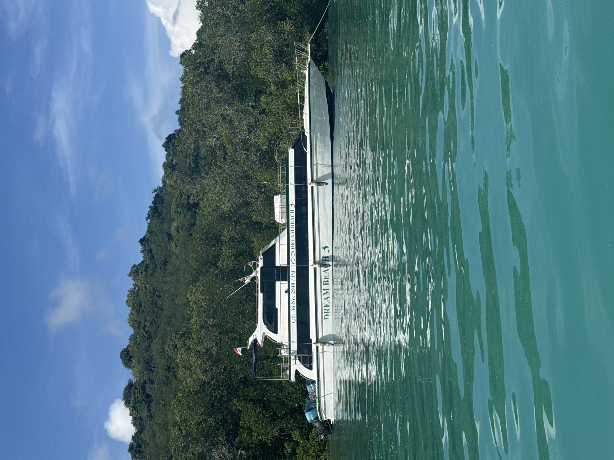 A horizontally oriented eye-level shot shows a motorboat, labeled "Dream Beach 3" floating on water. A forested hill is on the left and a mostly clear, blue sky with a few clouds is in the upper left. The image was taken in Nusa Penida, Indonesia.