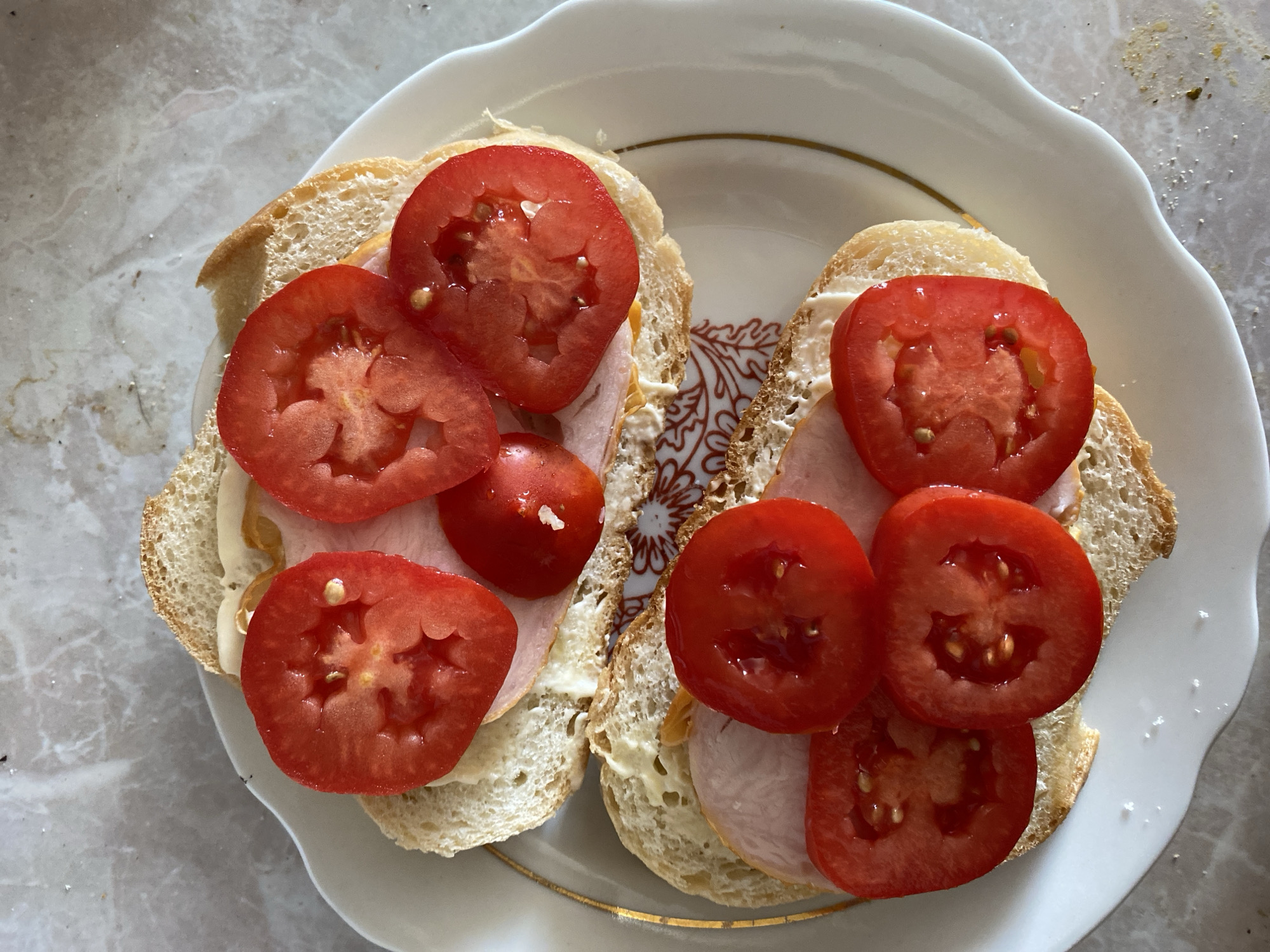 Two open-faced sandwiches are presented on a white plate with a gold rim. The sandwiches are made with sliced bread topped with mayonnaise, slices of processed meat, and slices of ripe red tomato. The plate sits on a marbled countertop. The scene appears to be indoors, likely in a kitchen or dining area, within the city of Chornomorsk, Ukraine.