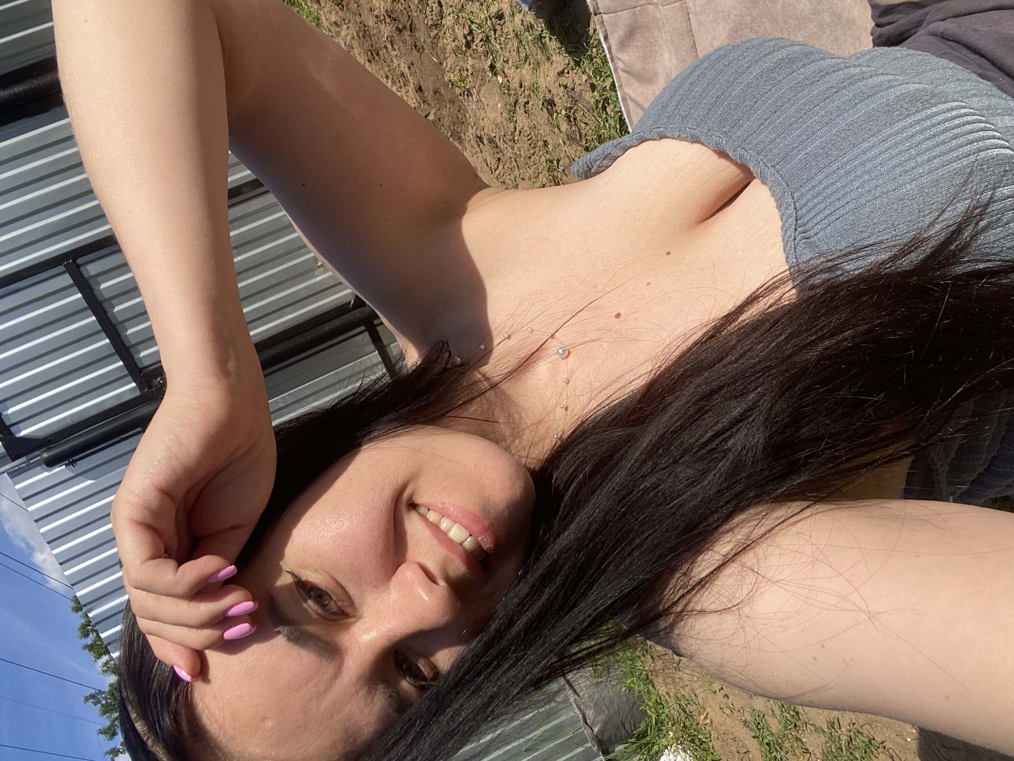 A female individual is depicted in a brightly lit outdoor setting, presenting a selfie-style shot. She possesses long, dark hair and has a fair to medium complexion, with visible small moles on her chest. Her left hand, featuring pink nail polish, is positioned near her forehead. She is smiling, revealing her teeth. The subject wears a light blue/grey ribbed tank top. A delicate necklace with small, round, metallic beads is visible on her décolletage. The background features a corrugated metal fence under a clear blue sky with minimal cloud cover, and some green foliage is visible above the fence on the left. The foreground consists of dry earth with sparse green vegetation, and a dark, possibly padded, surface is present below the subject. This scene is situated in the city of Chaikovsky, Russia.