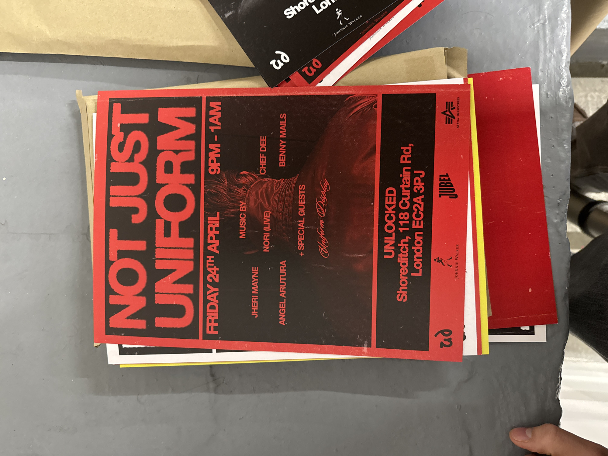 This media file captures a stack of promotional flyers resting on a grey, textured surface, possibly a table or counter. A person's hand is partially visible in the bottom right corner. The most prominent item is a red and black flyer for an event titled "NOT JUST UNIFORM".
The flyer provides details for a music event scheduled for FRIDAY 24TH APRIL, from 9PM - 1AM. It lists several performers and DJs including JHERI MAYNE, NORI (LIVE), ANGEL ARUTURA, CHEF DEE, and BENNY MAILS, with additional "SPECIAL GUESTS" and a "Uniform Display". The event is located at UNLOCKED Shoreditch, 118 Curtain Rd, London EC2A 3PJ. This location cue places the event in London, United Kingdom.
Sponsor logos visible on the flyer include ALPHA INDUSTRIES (top right), JUBEL (below location details), and JOHNNIE WALKER (bottom right and on other visible flyers). A stylized logo, possibly "ud" or "p2", appears at the bottom left. The flyer's design incorporates a dark, blurred image of what appears to be a person's head and torso, contributing to a dynamic, possibly edgy, aesthetic.
Beneath the main flyer, other promotional materials are visible, including red, yellow, black, and white cards, some also featuring the Johnnie Walker brand and the "ud" logo, suggesting they are related to the same event or a broader marketing campaign. A brown paper item, possibly an envelope or packaging, is partially visible in the top left corner. The scene depicts event publicity materials ready for distribution or display.
