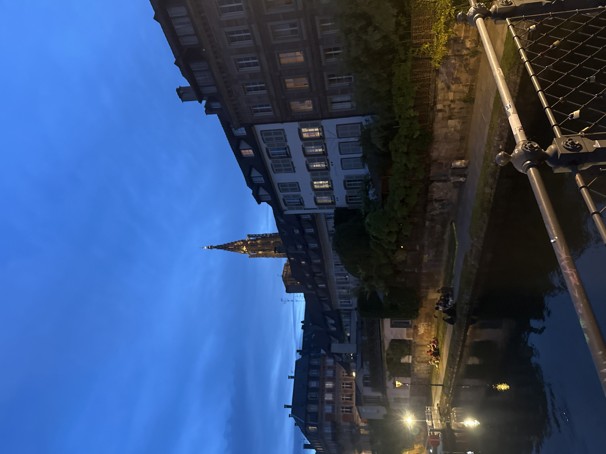 The image captures an evening scene in Strasbourg, France, depicting a canal or river lined with traditional European buildings under a deep blue twilight sky. The prominent spire of the Strasbourg Cathedral, illuminated against the fading light, rises in the mid-ground.
Along the left side of the waterway, multi-story buildings with numerous windows, some displaying warm interior lights, create a sense of urban life. The right bank features a pedestrian path bordered by lush trees and shrubs. Several groups of people are visible on this bank, appearing to be sitting or standing, enjoying the tranquil evening by the water.
In the immediate foreground on the right, the dark metal railing of a bridge is prominent. Many small "love locks" are attached to its mesh, a common sight on romantic bridges. Artificial lights from streetlamps illuminate parts of the scene and cast bright reflections on the dark surface of the water. The overall atmosphere is peaceful and serene. Faint, unreadable graffiti is visible on the canal wall, and small, unidentifiable stickers are present on the bridge supports.
