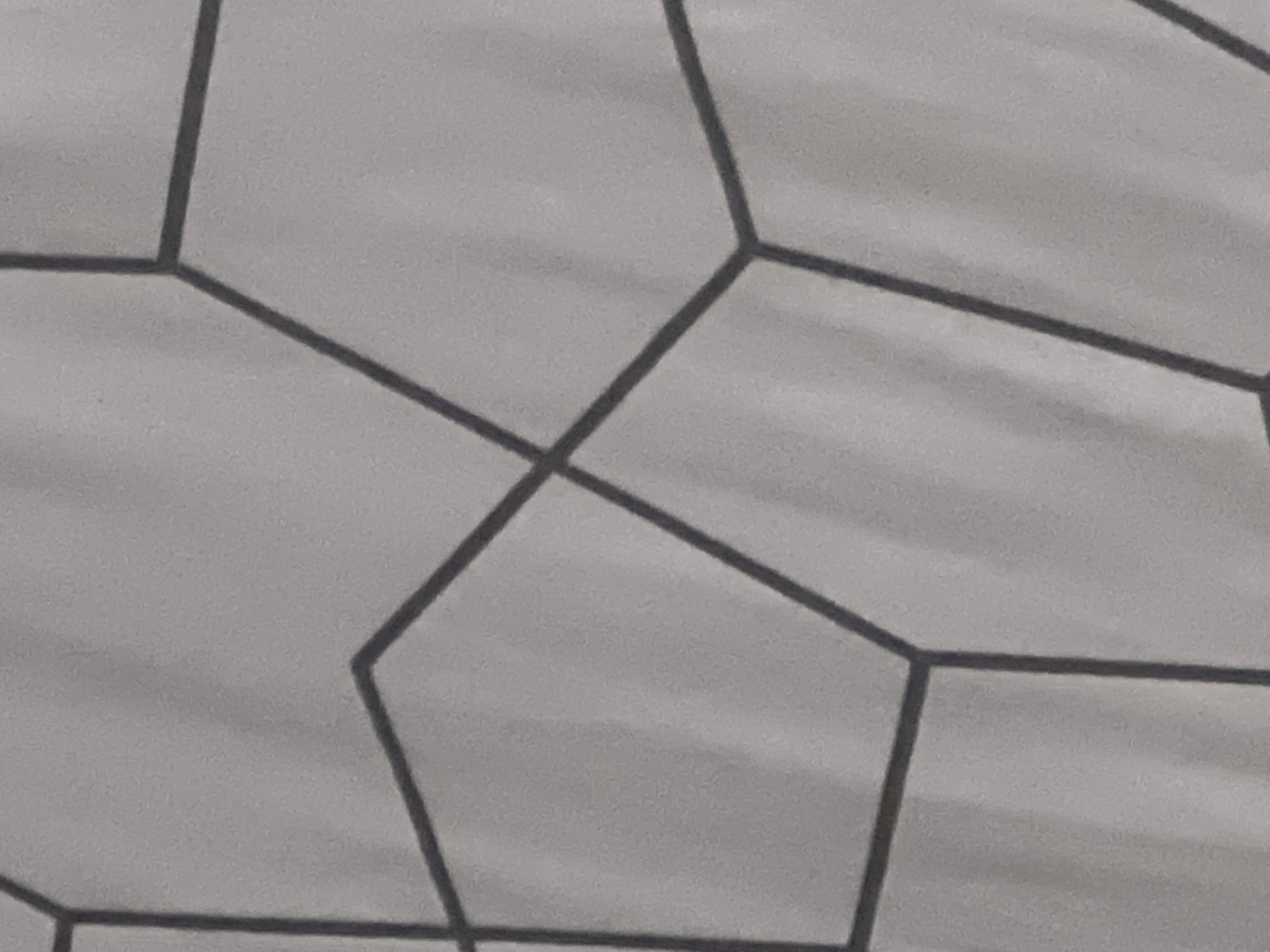 The image presents a close-up, overhead view of a light grey surface patterned with a network of dark grey to black lines. The lines form an irregular tessellation of geometric shapes, primarily identifiable as hexagons and pentagons, with several incomplete polygons extending beyond the frame's edges. The background exhibits subtle, uneven shading and textural variations across its light grey tone, suggesting a material like paper, fabric, or a painted wall. The lines themselves are not perfectly uniform in thickness or straightness, indicating either a hand-drawn origin or a printed design with textural elements. There are no discernible people, specific objects other than the lines and surface, or external environmental features.