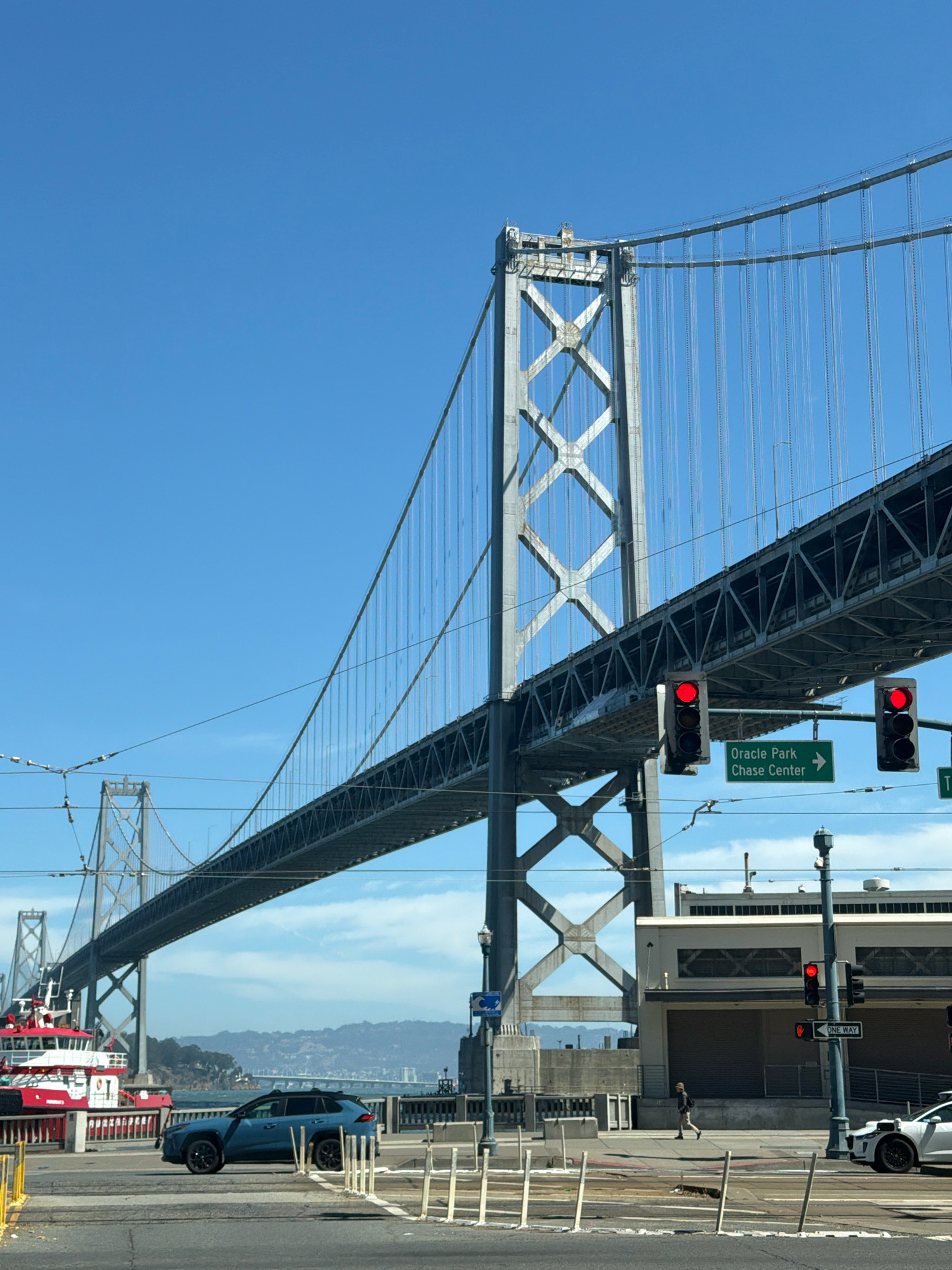 The image depicts a view of the San Francisco–Oakland Bay Bridge under a clear blue sky. The foreground features a blue SUV parked on a street, and to its left, a red and white boat is docked. Traffic lights are visible, with one showing red. A green sign indicates "Oracle Park Chase Center" with an arrow pointing right. In the background, mountains and a hazy cityscape can be seen across the water. A person is walking on the sidewalk in the middle ground.