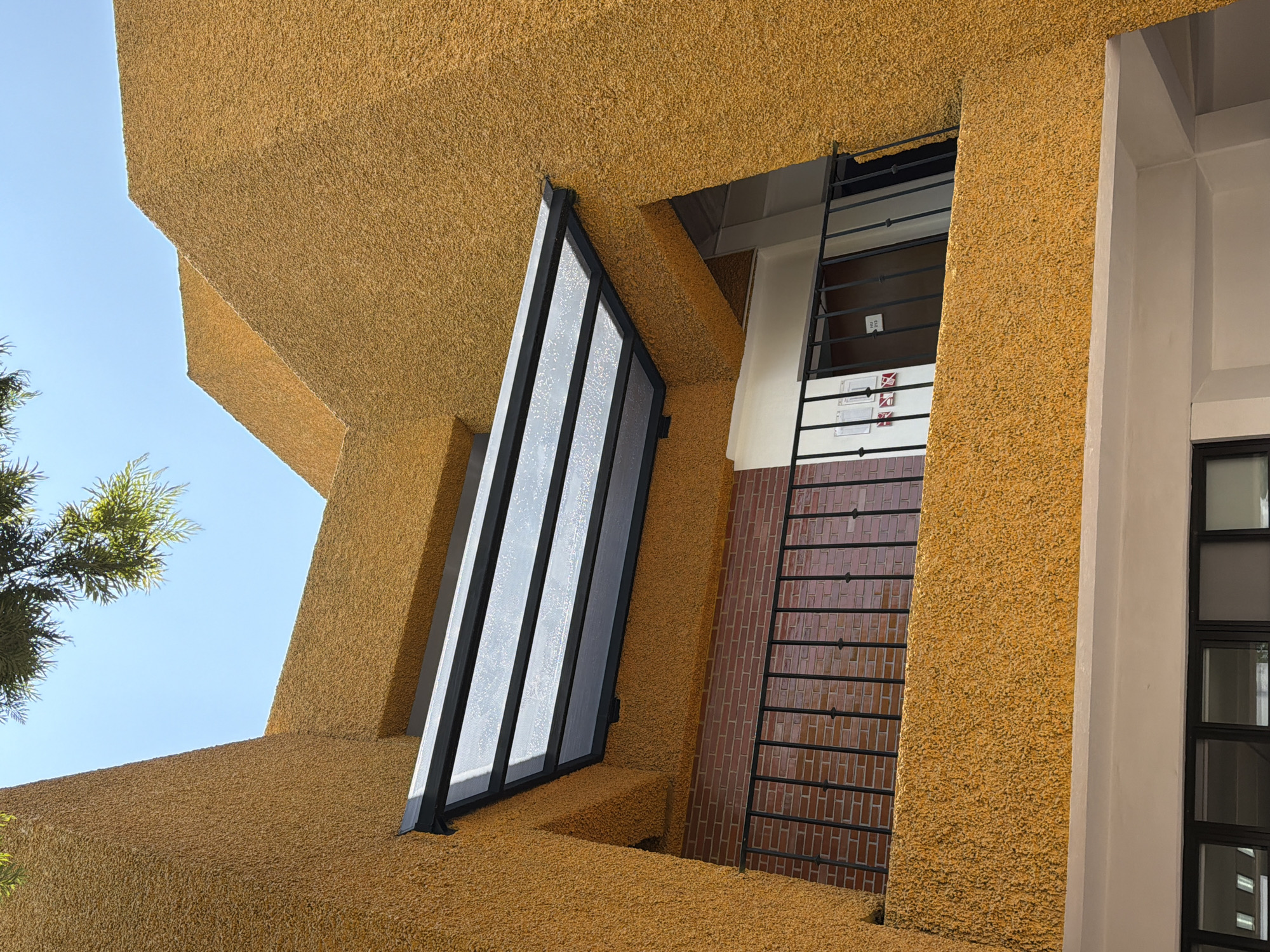 A low-angle, full-day exterior shot captures a building in San Andrés Cholula, Mexico. The building is predominantly textured in a coarse, mustard-yellow material. A large, black-framed glass structure protrudes diagonally from the building's façade. Adjacent to it is a window covered with a black metal grill, set against a background of dark red brickwork. To the right, a white column supports the building, with a visible black-framed window beside it. A clear, blue sky is visible in the upper-left corner, and some foliage is also present.
