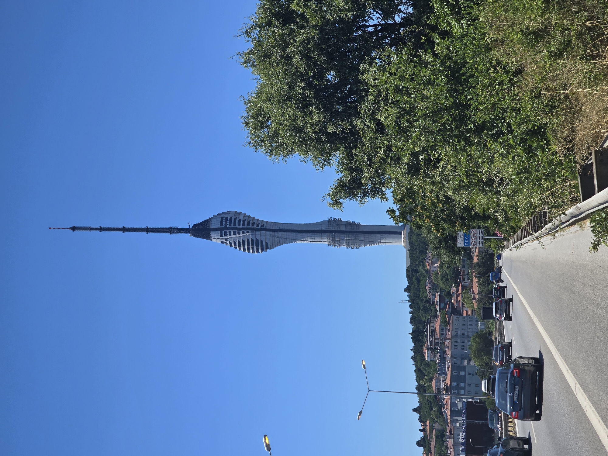 An eye-level, outdoor, wide shot captures a view of the Çamlıca Tower and surrounding cityscape on a clear, bright day. The modern, twisting tower dominates the right side of the frame, its metallic structure contrasting with the deep blue sky. Below the tower, residential buildings with red-tiled roofs are nestled on a green, tree-covered hillside. In the foreground, a multi-lane highway stretches into the distance, with several cars driving on it. A dense green tree canopy partially obscures the view of the tower on the right. The environment appears to be a developed urban area.