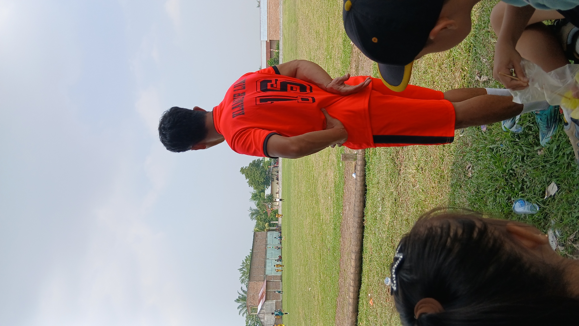 An outdoor, medium shot captures a group of people at a soccer field in Unknown, Indonesia. In the foreground, two children are seated on the grass, facing away from the camera. One child, on the right, is wearing a black baseball cap with a yellow brim and is holding a bag of snacks. The other child, on the left, has dark hair pulled back with a sparkly accessory.
Behind the children, a man wearing a bright orange soccer jersey with black trim and the number "15" on the back stands with his hands on his hips, looking towards a soccer game taking place in the distance. The field is a lush green grass, with scattered trees and buildings visible in the background. The sky is overcast. The scene appears to be a casual sporting event, with spectators and players present.