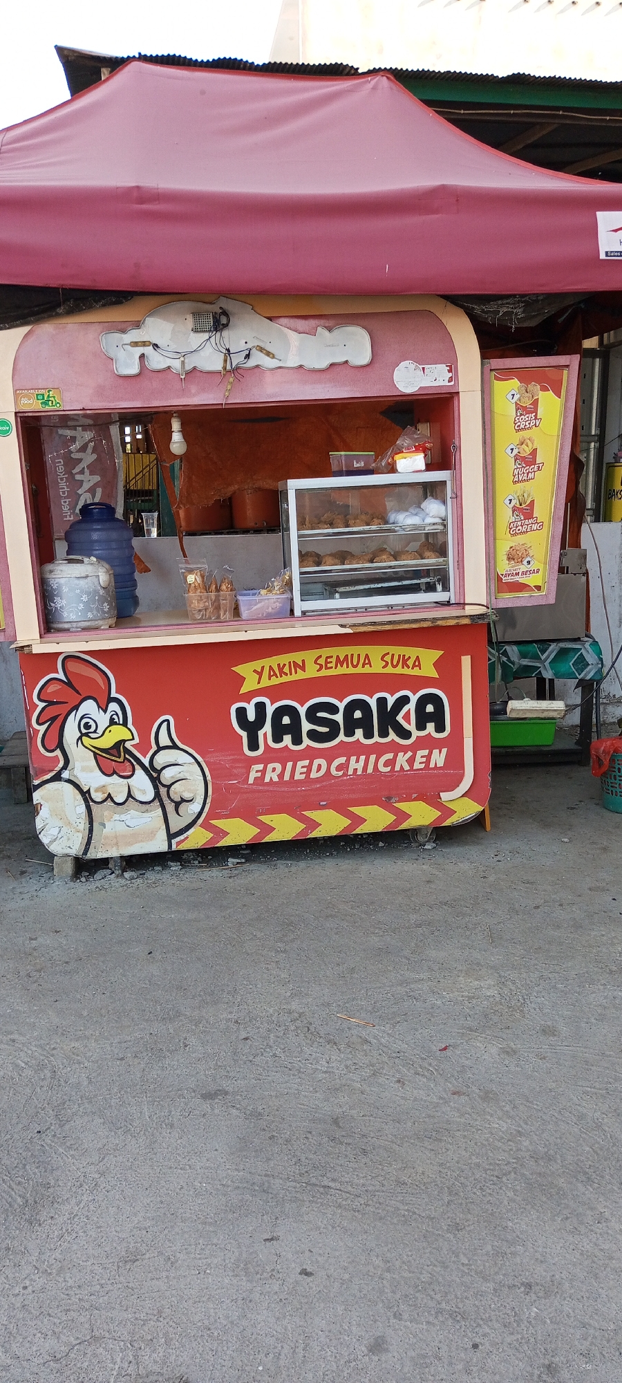This is a medium shot, eye-level photograph of a food stall. The stall is painted red and features a cartoon chicken giving a thumbs up. The stall's name, "YASAKA FRIED CHICKEN", is prominently displayed in large letters. Above the chicken, a yellow banner reads "YAKIN SEMUA SUKA" (roughly translated to "Everyone is sure to like"). Inside the stall, a glass display case shows fried chicken. Behind the counter, there are bags of snacks, a water dispenser, and cooking pots. The stall is set up on a concrete surface, and a red awning covers the top. The background shows a building with a corrugated metal roof and some wooden structures. This scene is located in Pakkanna, Indonesia.