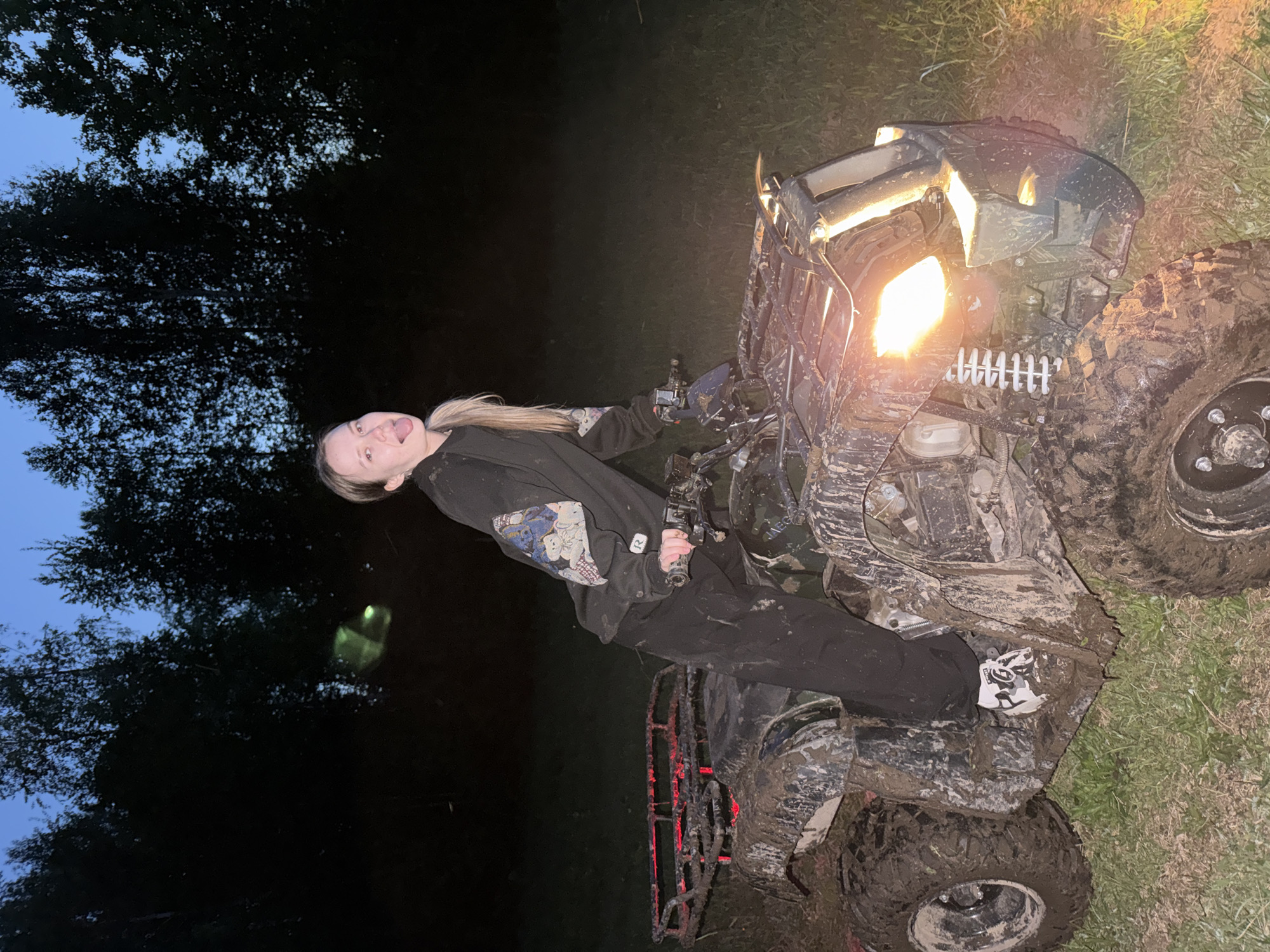 A young, blonde woman is sitting on a mud-covered ATV in an outdoor environment. The ATV's headlights are on, illuminating the immediate area. The background consists of trees and dark sky, indicating it is either dusk or night. The woman is sticking her tongue out and looking at the camera. Her clothes appear to be dark in color and also covered in mud. The location is unspecified beyond being an outdoor area likely in Zhigalki, Russia.
