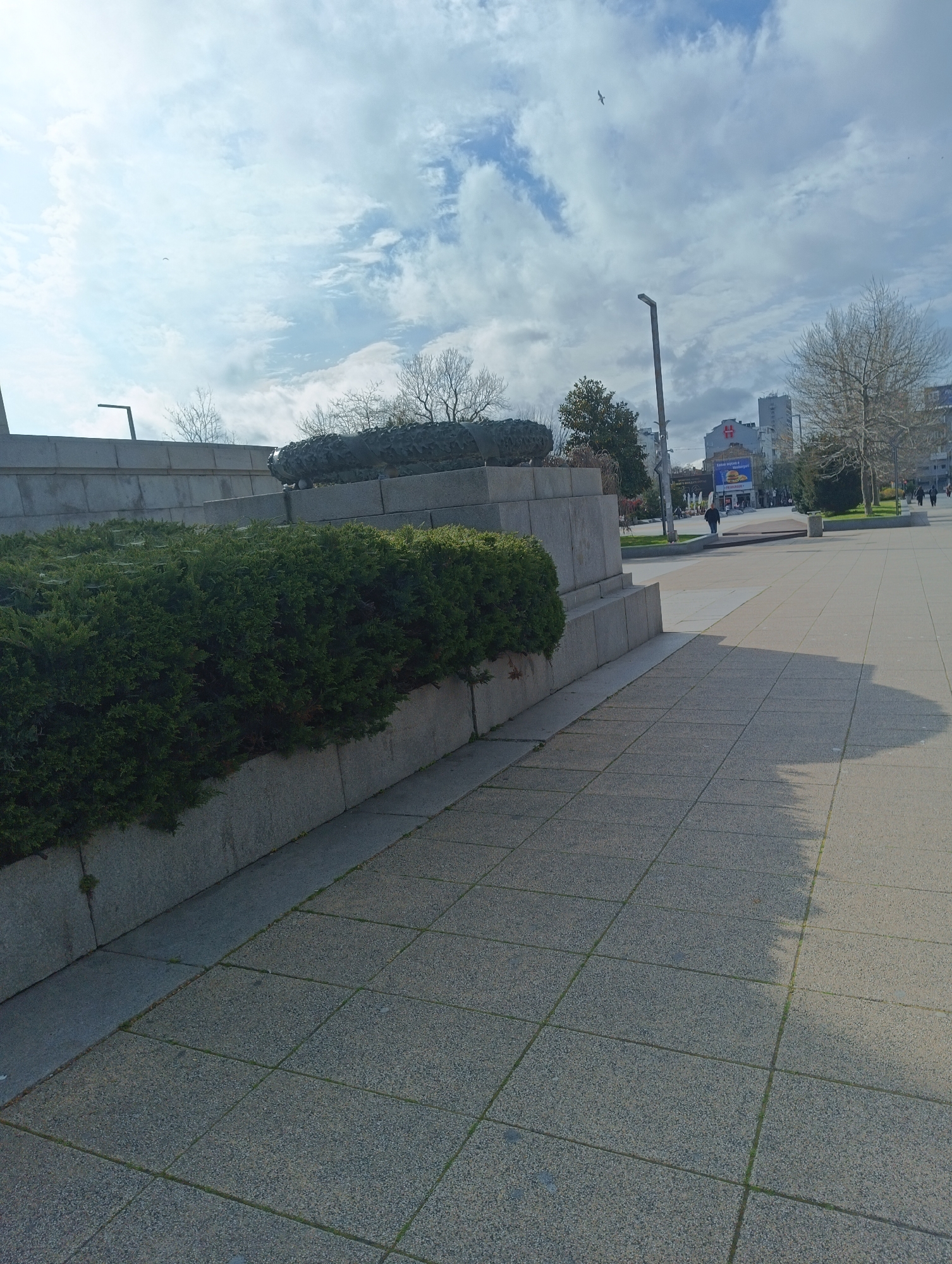 The image depicts a monument with evergreen bushes on the left, and a stone-paved walkway leading to a distant cityscape under a partly cloudy sky. A streetlight and bare trees are visible in the background. There are a few people walking in the distance. Based on visual cues, the location is likely in Burgas, Bulgaria, possibly near a central plaza or park.