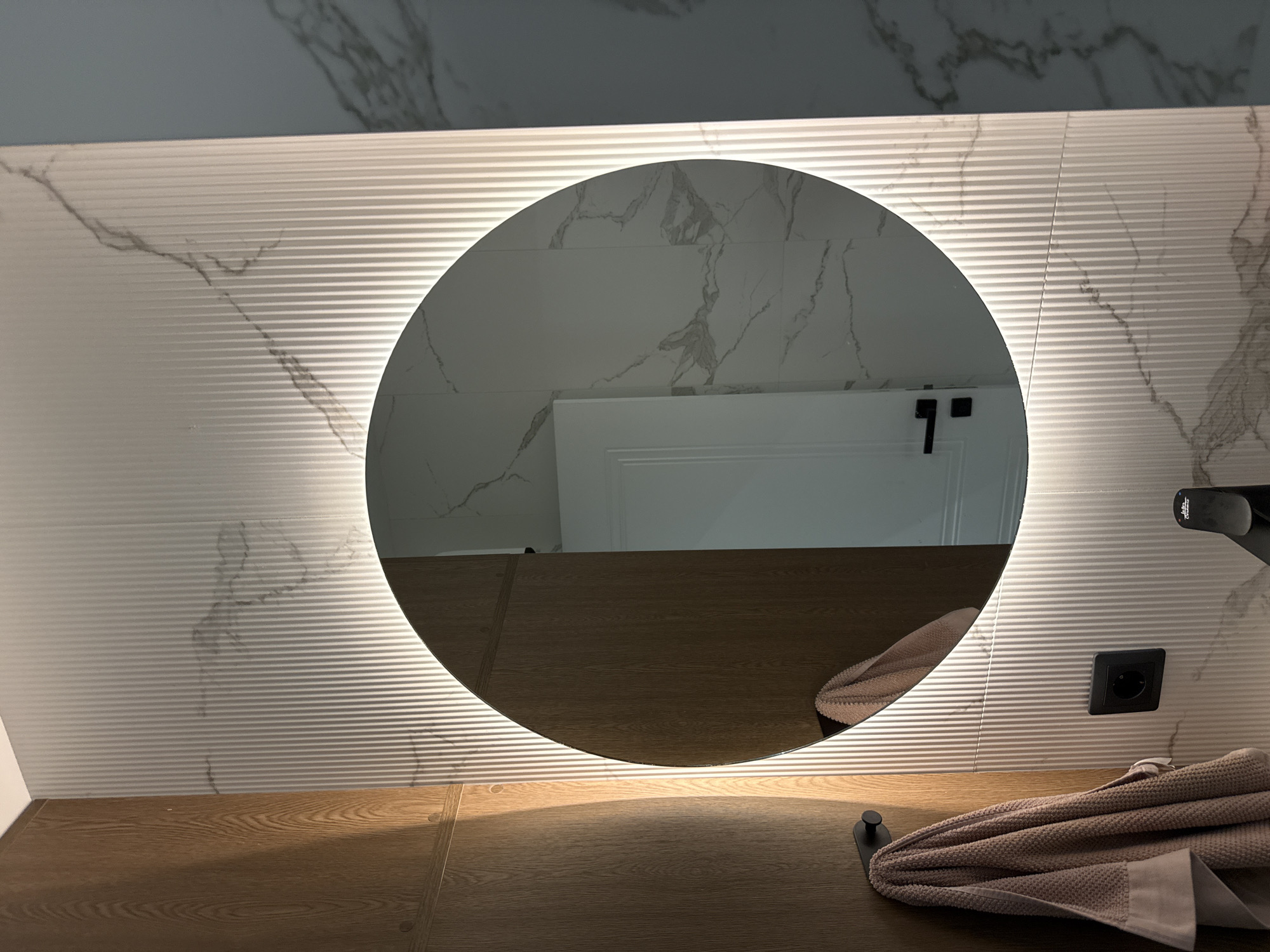 The image shows a bathroom interior. A round, illuminated mirror is the focal point, reflecting a portion of the bathroom. The wall behind the mirror is tiled with a white, horizontally-ridged tile featuring marble-like veins. Below the mirror is a wooden countertop. To the right of the mirror, a black faucet is visible. A folded towel lies on the countertop near a black towel hook. An electrical outlet is seen on the wall to the right of the mirror. The reflection in the mirror shows a white door with a black handle.
While the setting is a bathroom, there is no information in the image to suggest a location in Plovdiv, Bulgaria, or any other specific location.