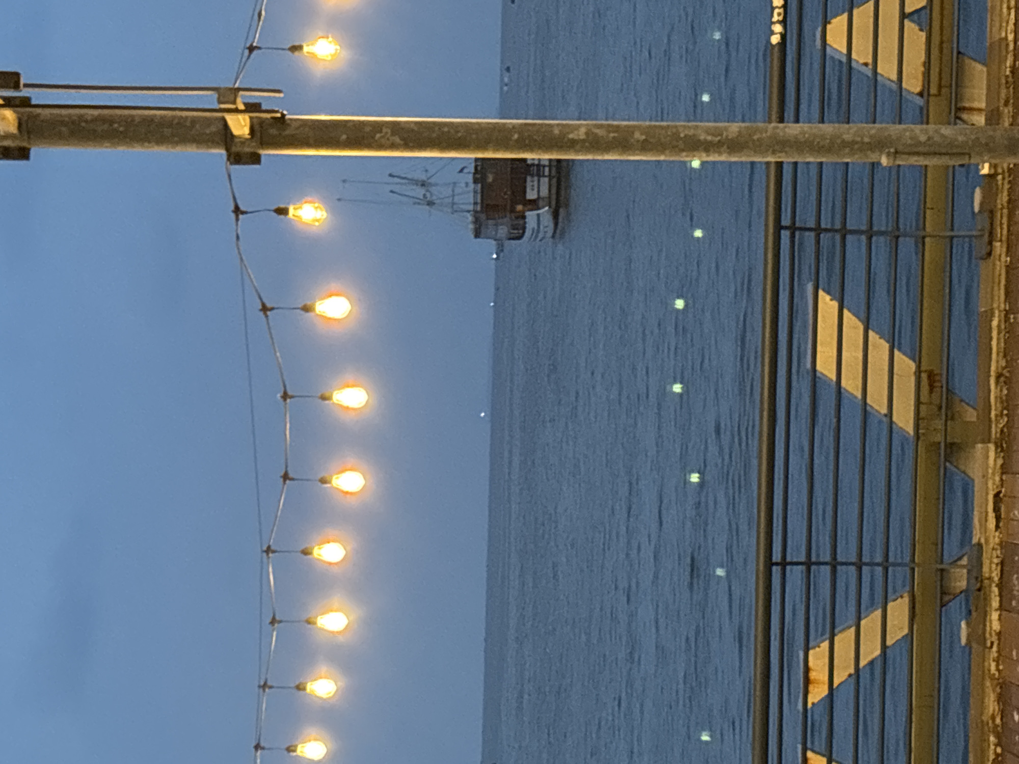 The image depicts a scene at dusk or dawn, with a body of water (possibly the sea) filling the majority of the frame. A vintage-style ship is visible in the distance on the water. A string of glowing lights is strung along a metallic horizontal support, running vertically on the left side of the frame. To the right of the lights, a barrier made of metal bars runs vertically along the right side of the frame. The barrier has a yellow decorative design on it. Based on the metadata, this location is most likely somewhere in the North Jakarta area of Indonesia.