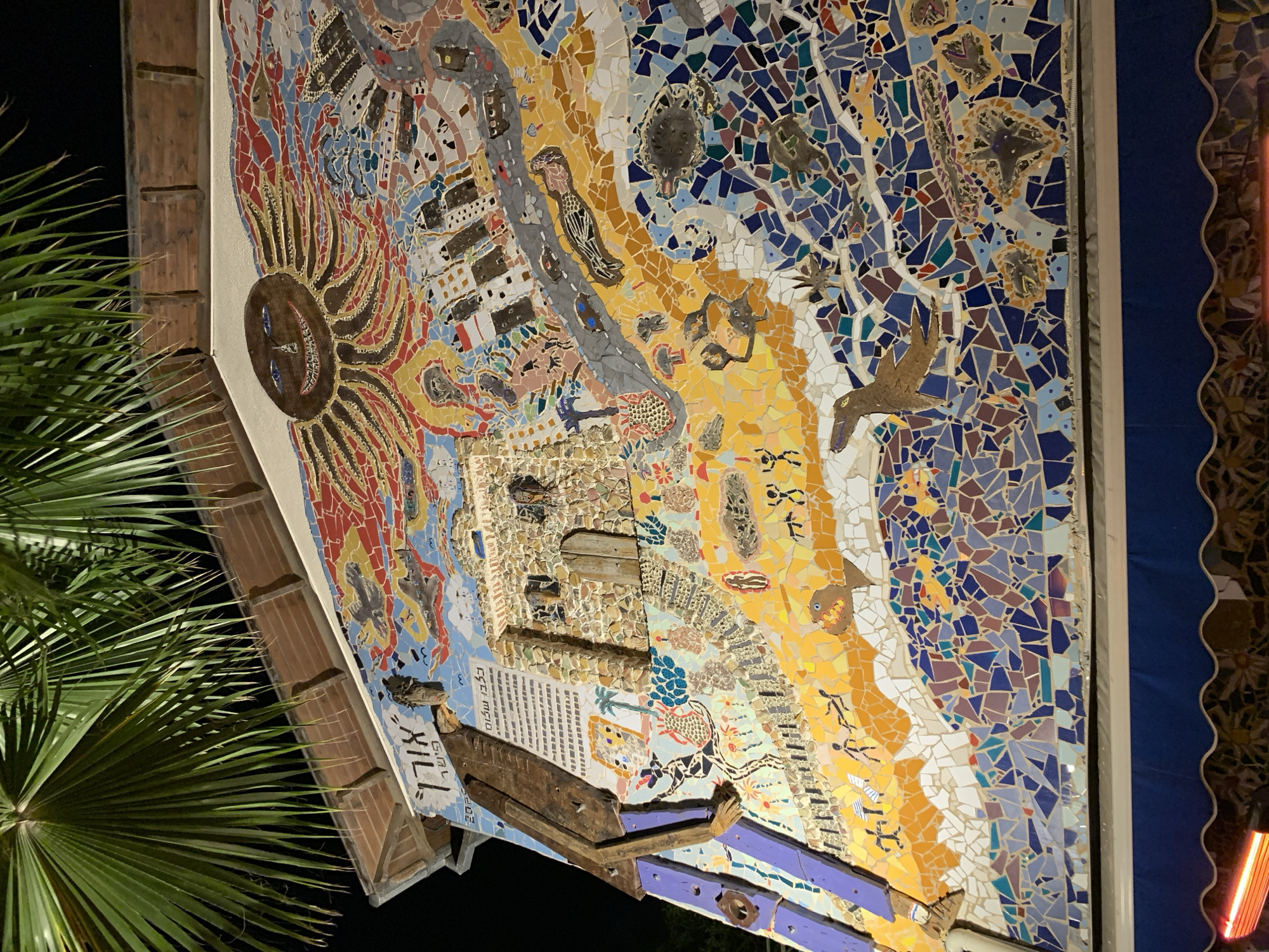 The image displays a large, complex mosaic mural adorning the exterior wall of a building at night in Tel-Aviv, Israel. Above the mural, against a dark sky, numerous dark green palm fronds hang into the frame, partially obscuring a brown-tiled roofline.
The mural itself is composed of countless small, irregularly shaped mosaic tiles and incorporates three-dimensional elements. In the upper central section, a large, stylized sun figure with a dark brown, smiling face and blue eyes radiates dark reddish-brown, tentacle-like rays across a sky depicted in light blue and white mosaic. To the left of the sun, Hebrew text "אורי פישל" (Uri Fishel) and the year "2023" are visible. Several dark-colored aircraft are depicted flying in the upper right sky area.
The central portion of the mural features a prominent, textured structure resembling an ancient stone building or fortress, complete with a small wooden door and several small window-like openings; this element slightly protrudes from the wall surface. To the left of this structure, a tall, slender, humanoid figure constructed from dark brown, jointed segments and purple lower limbs is affixed vertically to the building's edge. Behind the upper part of this figure, a white rectangular panel with dark text, likely Hebrew, is attached to the wall. To the right of the stone structure, a modern cityscape is depicted, including buildings, roads, and various vehicles like cars and buses.
Below these central elements, the mural transitions from a land depiction, characterized by a mosaic of sandy yellow and orange tones, to a water depiction using various shades of blue, purple, and white tiles. The land portion includes small, abstract human figures engaged in various postures, stylized plants, and a brown bird-like creature. The water portion features mosaic representations of marine life, including a seal-like creature, several sea turtles, and starfish.
At the bottom of the frame, a dark blue, scalloped awning is partially visible directly beneath the mosaic. A bright, elongated red light source is present at the very bottom left, below the awning.