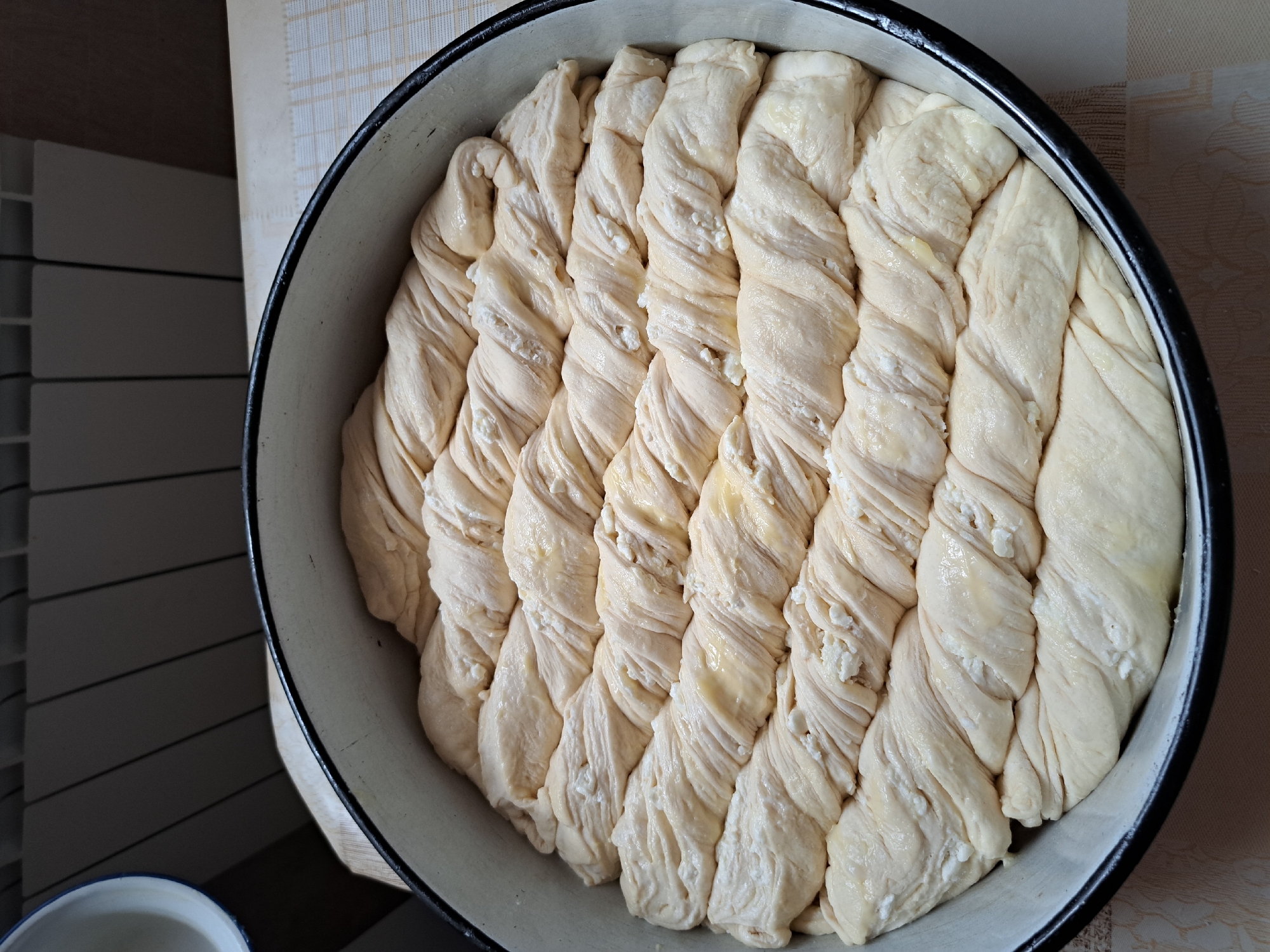 In a round, black-rimmed baking pan, a prepared dough is arranged in parallel, twisted ropes. The dough appears to be light in color and has a slightly oily sheen in places, with small white crumbles, possibly cheese or ricotta, visible between the twists. The pan is sitting on a pale, patterned tablecloth. In the background, a white bowl with a blue rim is partially visible on the left, and a white radiator with vertical slats is present on the upper right, against a dark wall. The lighting suggests an indoor setting, possibly a kitchen. The scene is devoid of people or any other animate subjects.