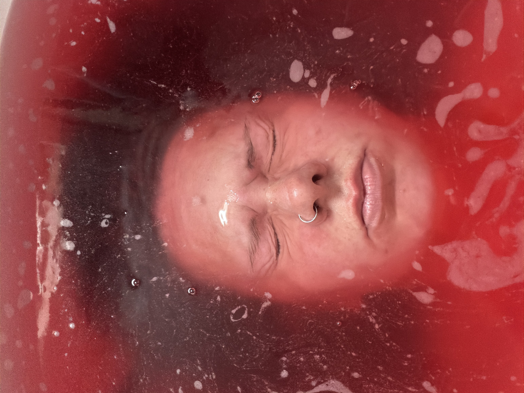 A person's face, eyes closed, floats in a red-tinted liquid, possibly water. A silver nose ring is visible. The environment is a liquid bath, with visible bubbles. There is no indication of this picture being taken in Chaikovsky, Russia.