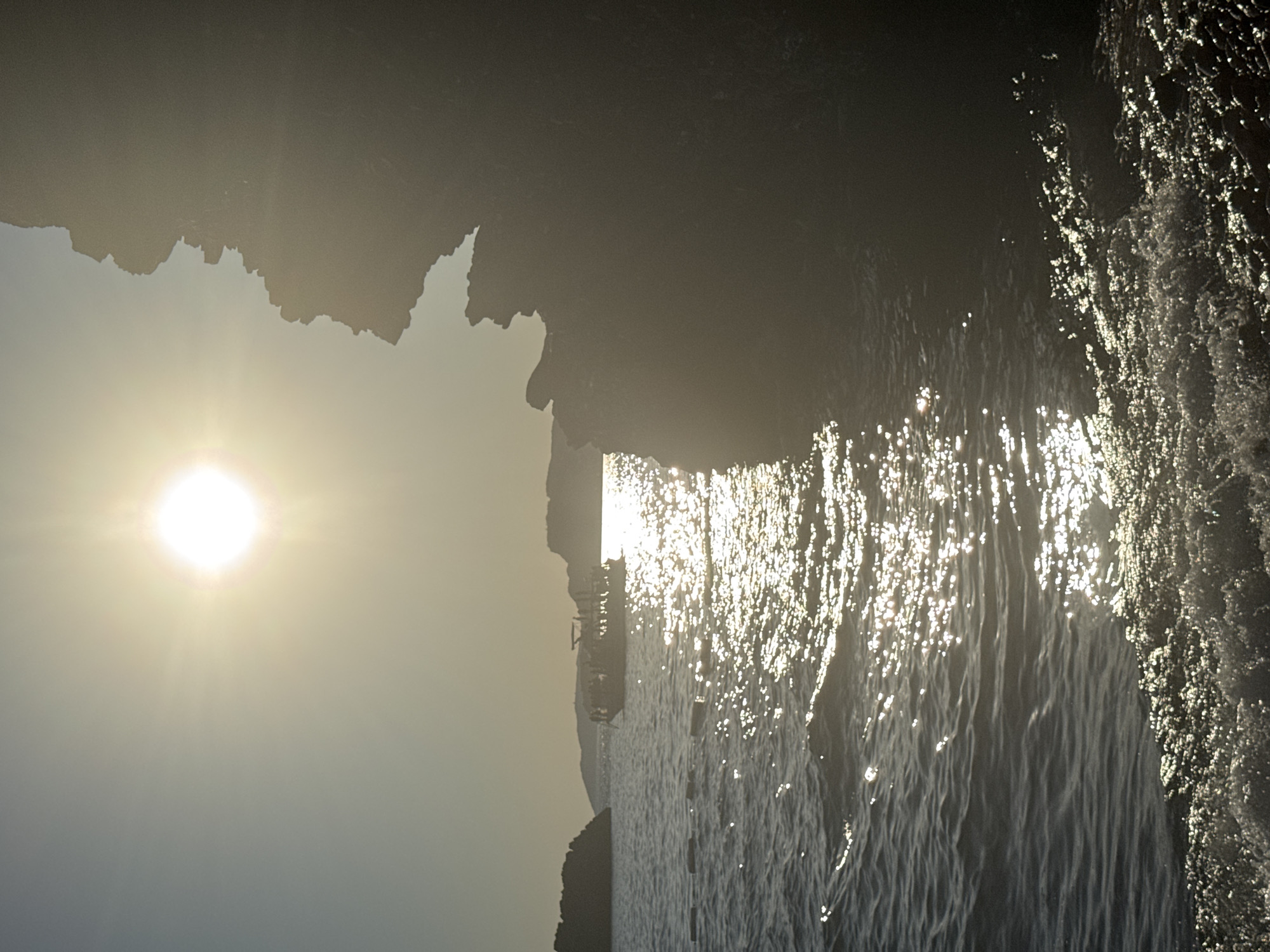 The image depicts a sunlit body of water with waves crashing against the shore, with a steep rock formation on the right side casting a shadow over the water. In the distance, a boat filled with people sails across the water. The sun is bright, creating a glistening effect on the water. The scene is set against a backdrop of a hazy sky and distant hills. The location is identified as Sveti Stefan, Montenegro.