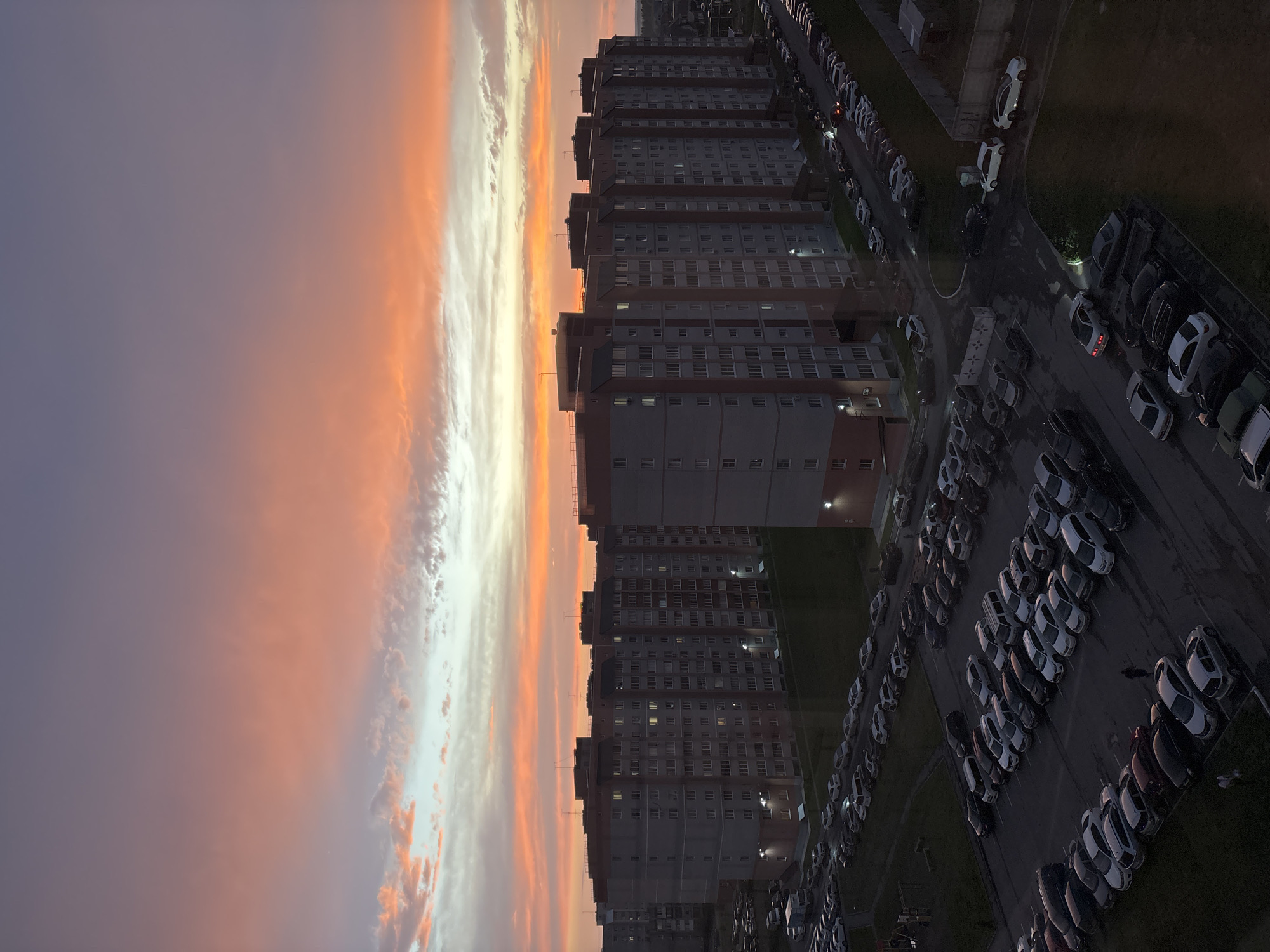 This is a vertical, high-angle, outdoor, photographic image of a residential area in Novoaltaysk, Russia, during a sunset. The sky is filled with vibrant orange, pink, and purple clouds. In the foreground, multiple apartment buildings, characterized by their grey facades and red accents, are visible. Rows of parked cars occupy the asphalt areas between and around the buildings. The lighting suggests it is dusk, with artificial lights illuminating some of the apartment windows and parking lot areas. The scene is quiet, with no apparent activity other than the static presence of buildings and vehicles.