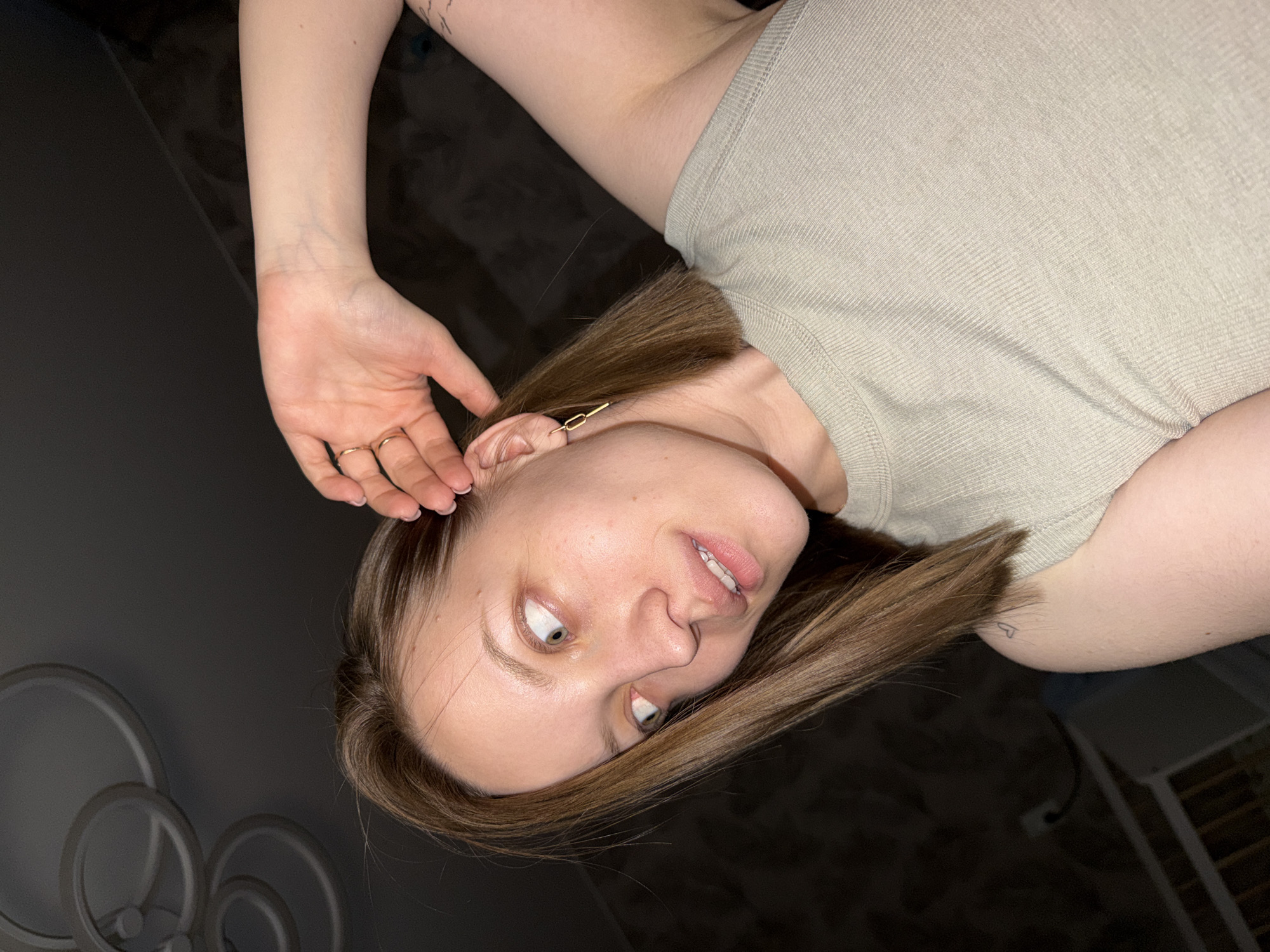 A woman with shoulder-length, brown hair lies on a dark, possibly upholstered surface. She wears a light beige shirt. Her left hand is near her ear, adorned with two rings. She has an earring in her left ear. Her eyes are directed upwards and to the left. In the lower left corner of the image, a light fixture composed of multiple circles is visible. A corner possibly a window or a shelf can be seen in the bottom right of the frame. The location is unknown and there is no evidence to indicate Reshetnikovo, Russia.