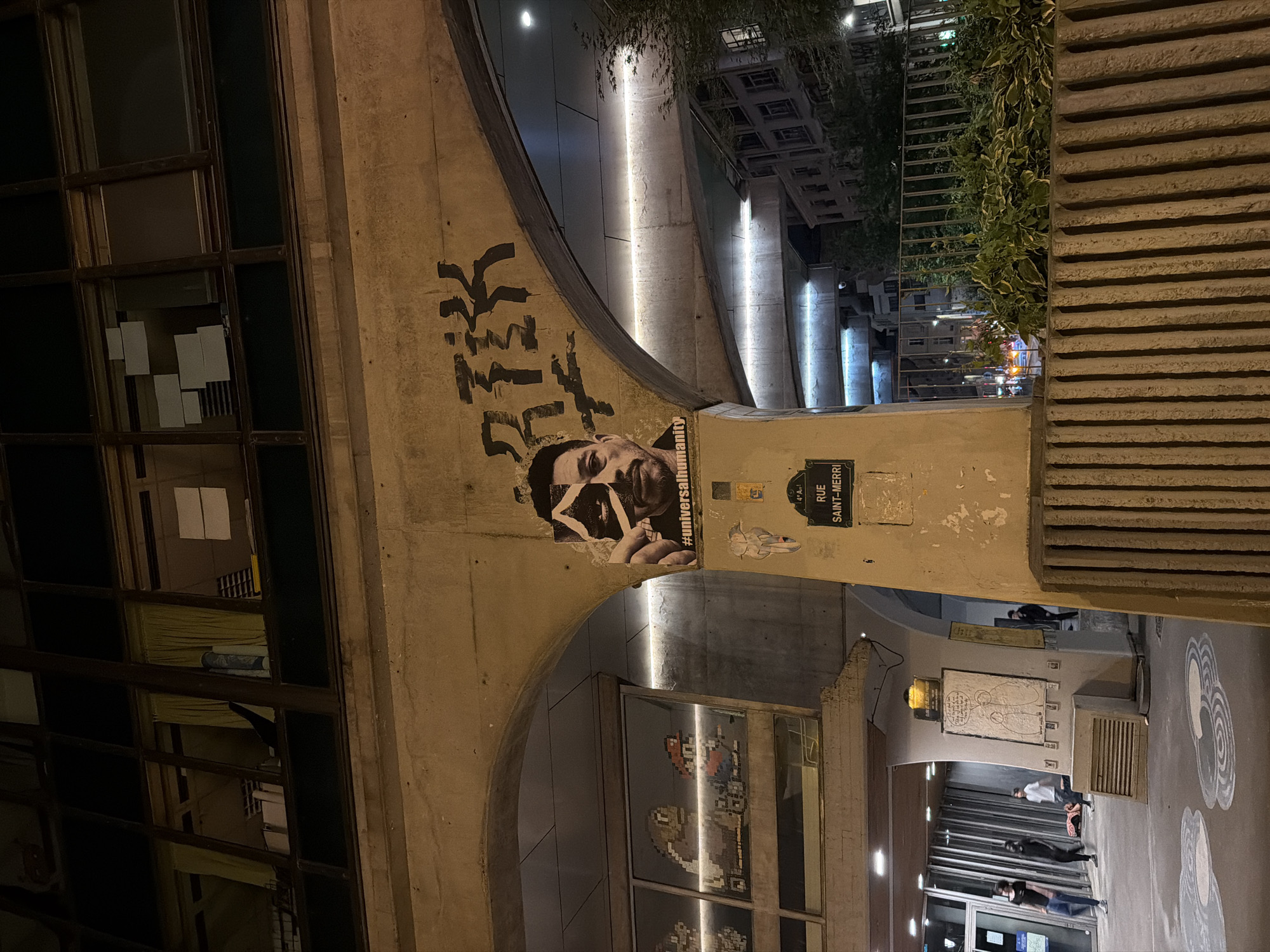 The image depicts a street scene in Paris, France at night. A concrete arched structure supports the street above, and underneath the arch, there are shopfronts and a walkway. The archway has graffiti, a poster of a person's face, and a street sign that reads "Rue Saint-Merri, 4e Arr." in French. People can be seen walking along the walkway. Above the arch, there are buildings, trees, and streetlights. There appears to be a pedestrian bridge that connects the buildings across the street above.