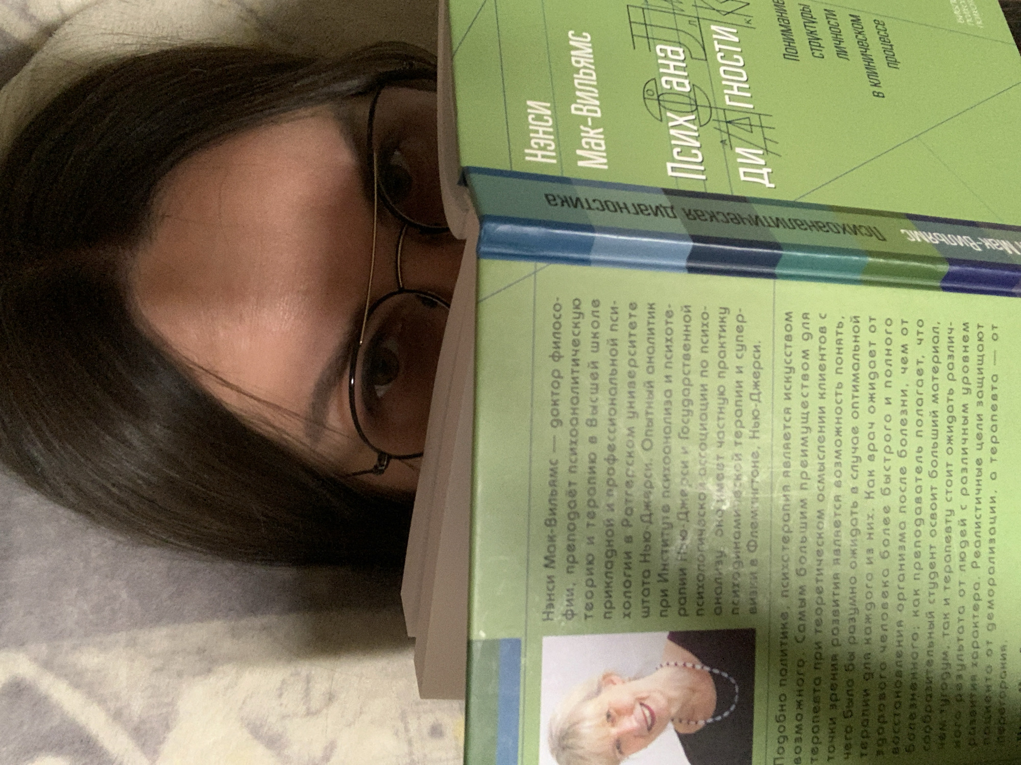 A close-up shot shows a person with dark hair and glasses looking at a book with a green cover. The book is open and the person's face is partially obscured by it. The text on the book's cover is visible, with the title "Психана/Диагностики" prominently displayed. The image appears to have been taken indoors, likely in a residential setting, given the soft lighting and casual nature of the scene. No specific location in the city of Unknown, Unknown can be determined from the image.