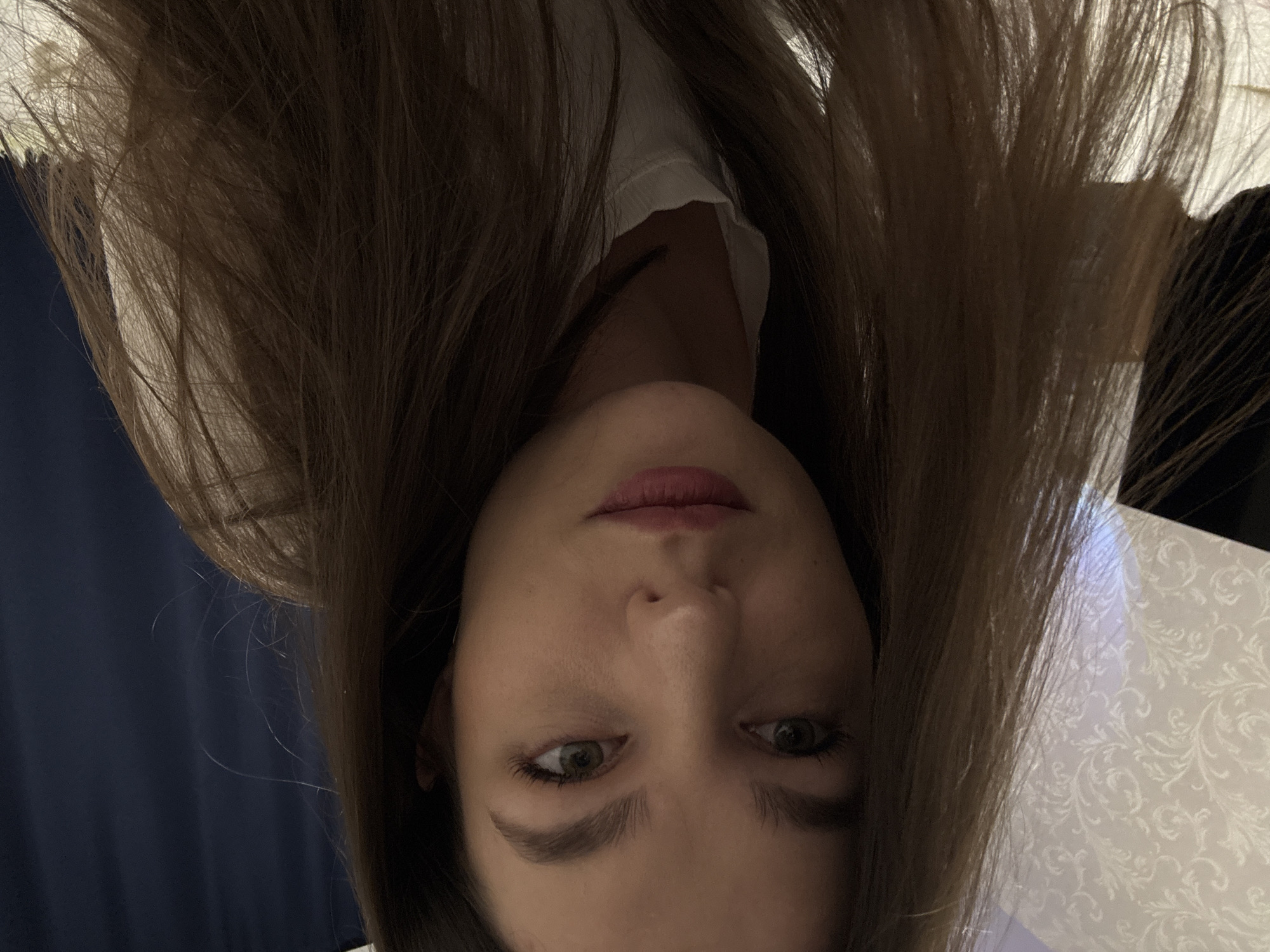 A woman with long brown hair is seen in a selfie taken from an upside-down perspective. Her face is framed by her hair. She has light skin, visible eyebrows, green eyes, and pink lips. She is wearing a white top with a ruffled neckline. The background appears to show a blue curtain and a patterned white surface. It is not possible to determine the location as Chaikovsky, Russia from the image content.