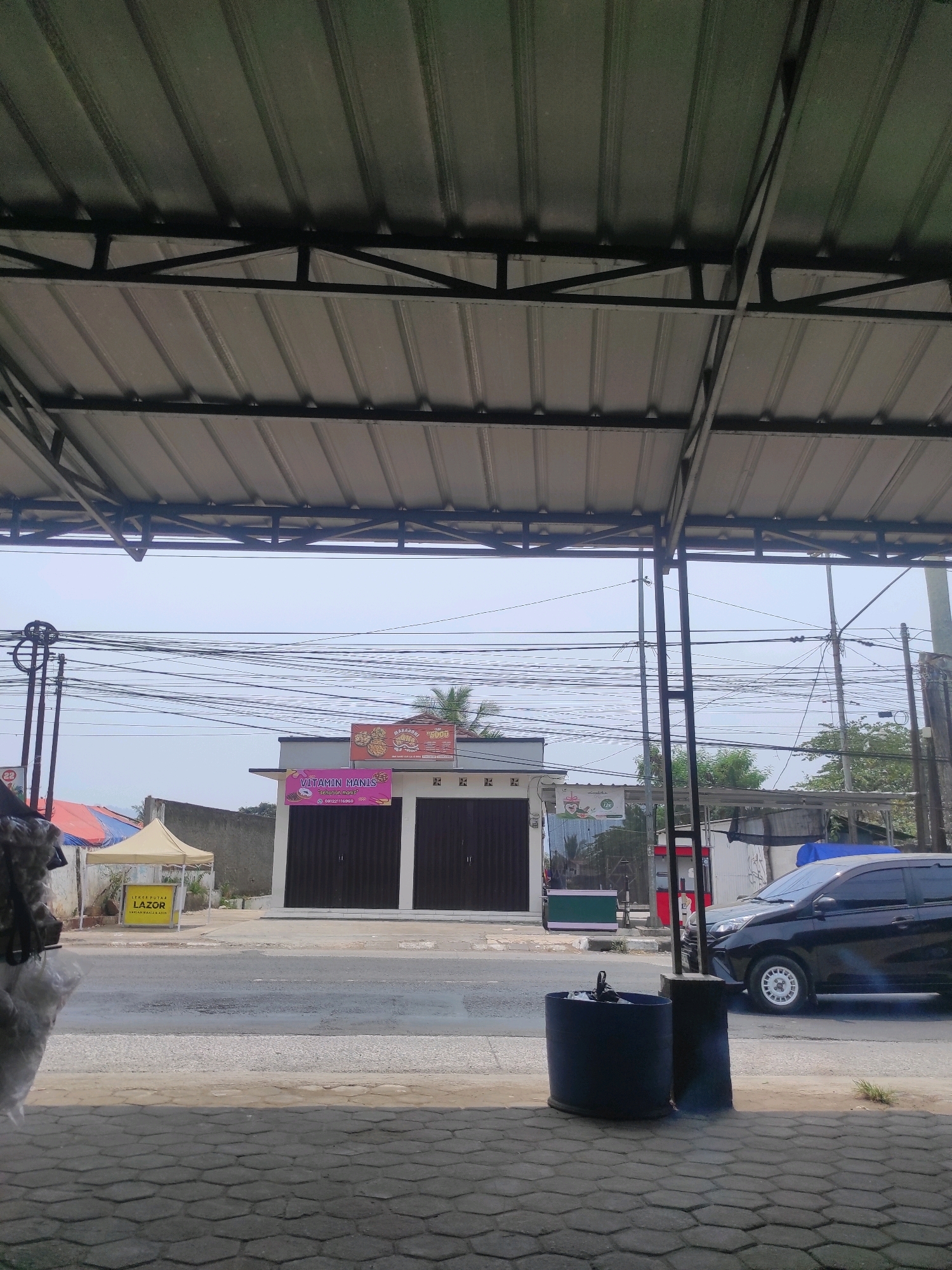 The image shows a view from under a metal awning, looking out onto a street in Cipatat, Indonesia. A two-story building with dark roll-up doors is visible, featuring signs for "KECERIAAN" and "VITAMIN MANIS". To the left of the building, there are tents and a yellow sign with "LAZOR" written on it. A black car is parked on the right side of the street. Overhead, a complex network of power lines stretches across the sky. In the foreground, a large dark blue trash bin is situated on a paved area. The overall scene depicts a commercial street with a mix of businesses and infrastructure under a cloudy sky.