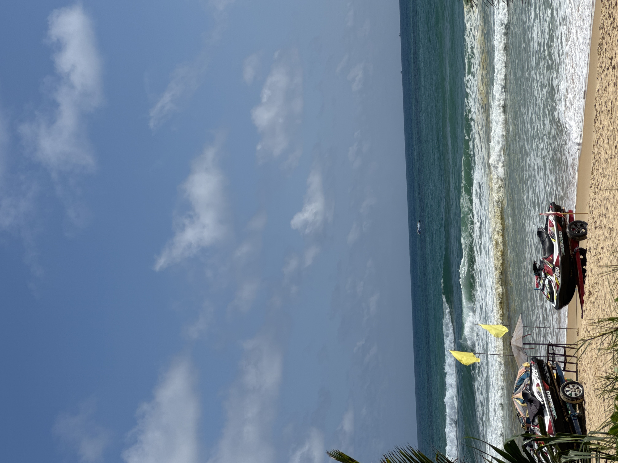 The image depicts a sunny day at Karon Beach in Karon, Thailand, with a clear blue sky dotted with scattered white clouds. The turquoise ocean stretches to the horizon, with moderate waves breaking onto the sandy shore. In the foreground, two jet skis are parked on trailers on the beach. One jet ski, mostly obscured by a cover, is on a trailer with a smaller trailer beside it. The second jet ski is fully visible and positioned to the right. Two yellow flags are planted in the sand between the jet skis and the water. Palm fronds are visible on the left side of the frame, and beach grass grows in the immediate foreground. In the distance, several small boats are visible on the water.