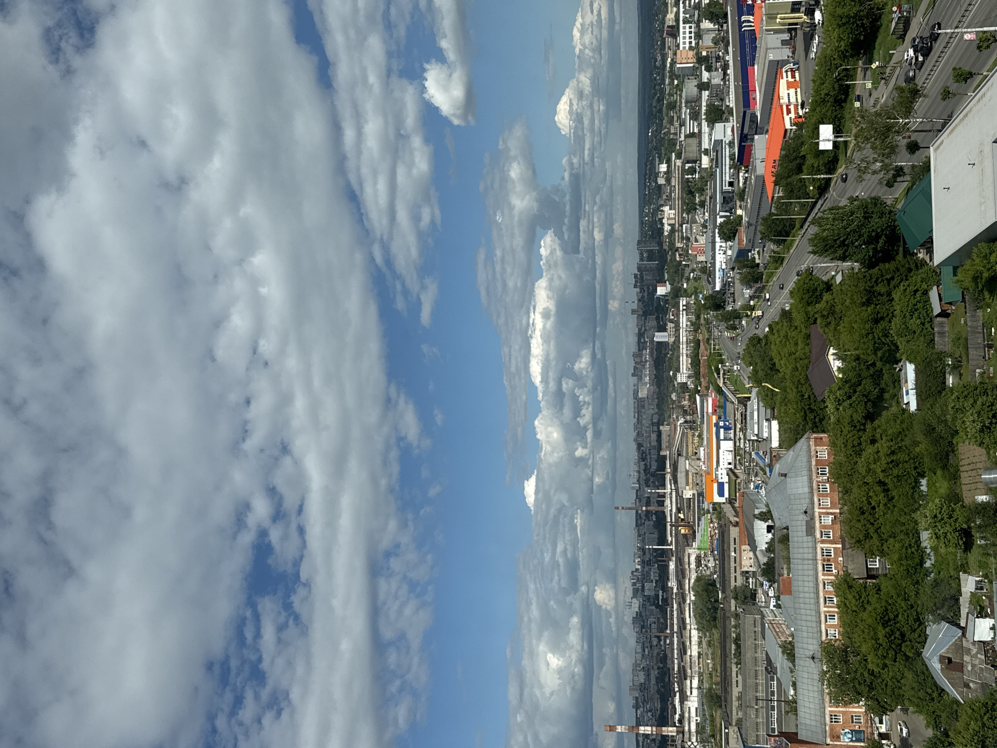 The image depicts an aerial view of an industrial area in Izhevsk, Russia, under a cloudy sky. In the foreground, there is a large, multi-story brick building with a gray metal roof, surrounded by dense green trees. Several smaller structures, including sheds and what appear to be garden plots, are also visible in the lower left portion of the frame.
A road with moderate traffic, including cars and buses, curves through the middle ground, bordered by more green trees. Beyond the road, a variety of industrial buildings are spread across the landscape, some with colorful facades (orange, blue, red, white), and others appearing as large, gray, utilitarian structures. Prominent features include several tall, striped smokestacks in the distance, characteristic of industrial facilities.
The background shows a cityscape with numerous buildings, suggesting the urban area extends beyond the industrial zone. The sky is filled with a mix of bright white cumulus clouds and patches of blue sky. The overall impression is one of a working industrial district on a partially overcast day.