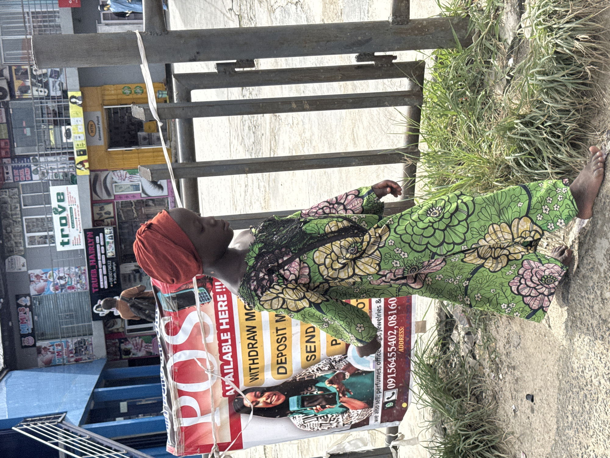 A woman in a green floral dress and red headwrap lies on a concrete surface beside grass, leaning against a metal railing and a POS advertising sign in Ado, Nigeria. The sign advertises money withdrawal and deposit services, along with contact numbers. The background includes storefronts and street scenes, suggesting a busy commercial area.