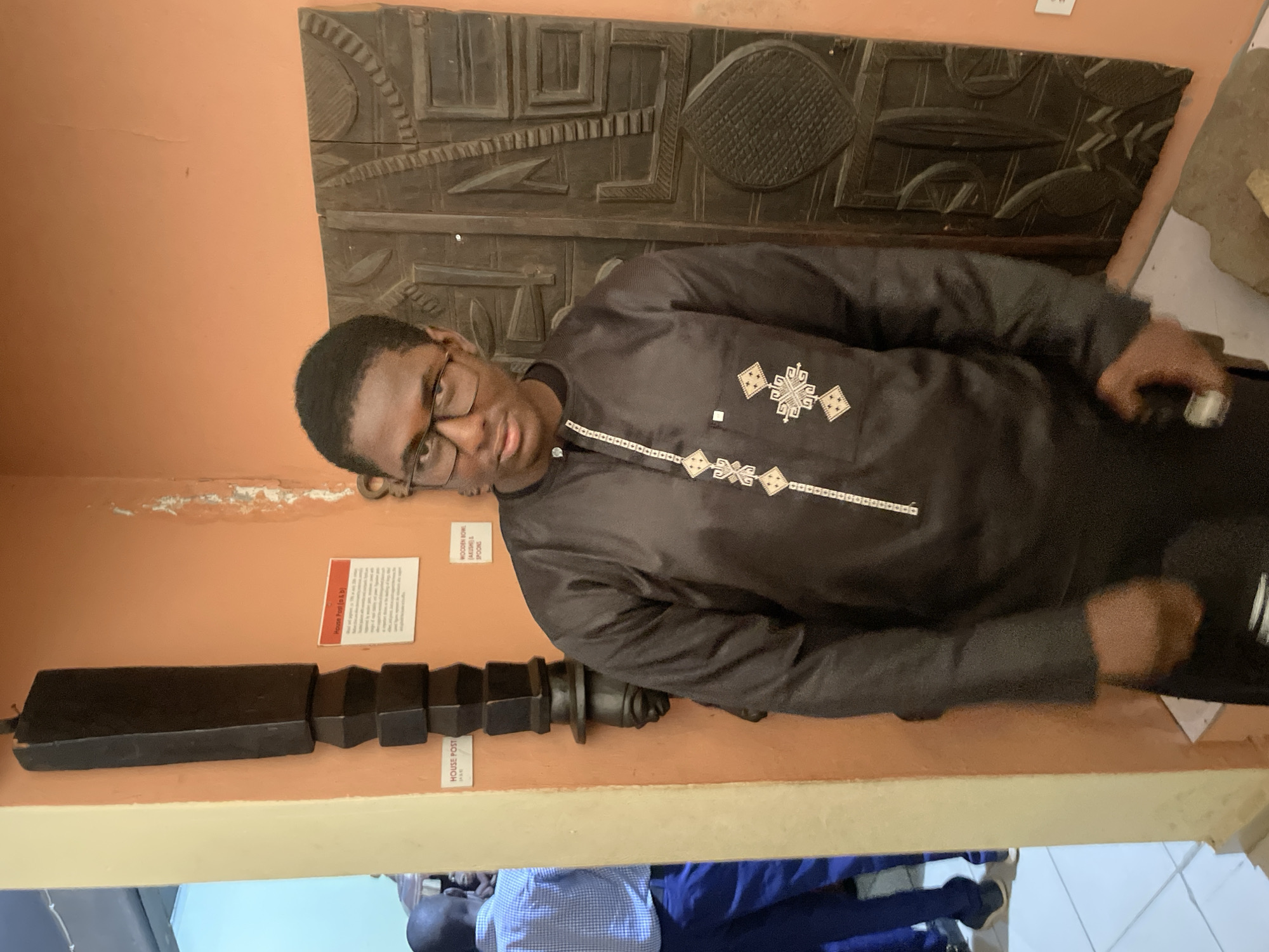 In Unguwar Shanu, Nigeria, a young Black male wearing glasses and a black traditional tunic stands in front of an orange wall. He is positioned slightly to the right of the frame. To his left, a tall, dark, carved wooden object, likely a house post, is mounted on the wall. Behind him, to his right, is a large, intricately carved wooden panel, also mounted on the wall. The wooden panel features geometric patterns and possibly figural elements. In the background, to the left of the house post, another person is visible, wearing a blue plaid shirt and blue trousers, facing away from the camera. The floor appears to be tiled. The overall scene suggests a museum or cultural exhibition space.
