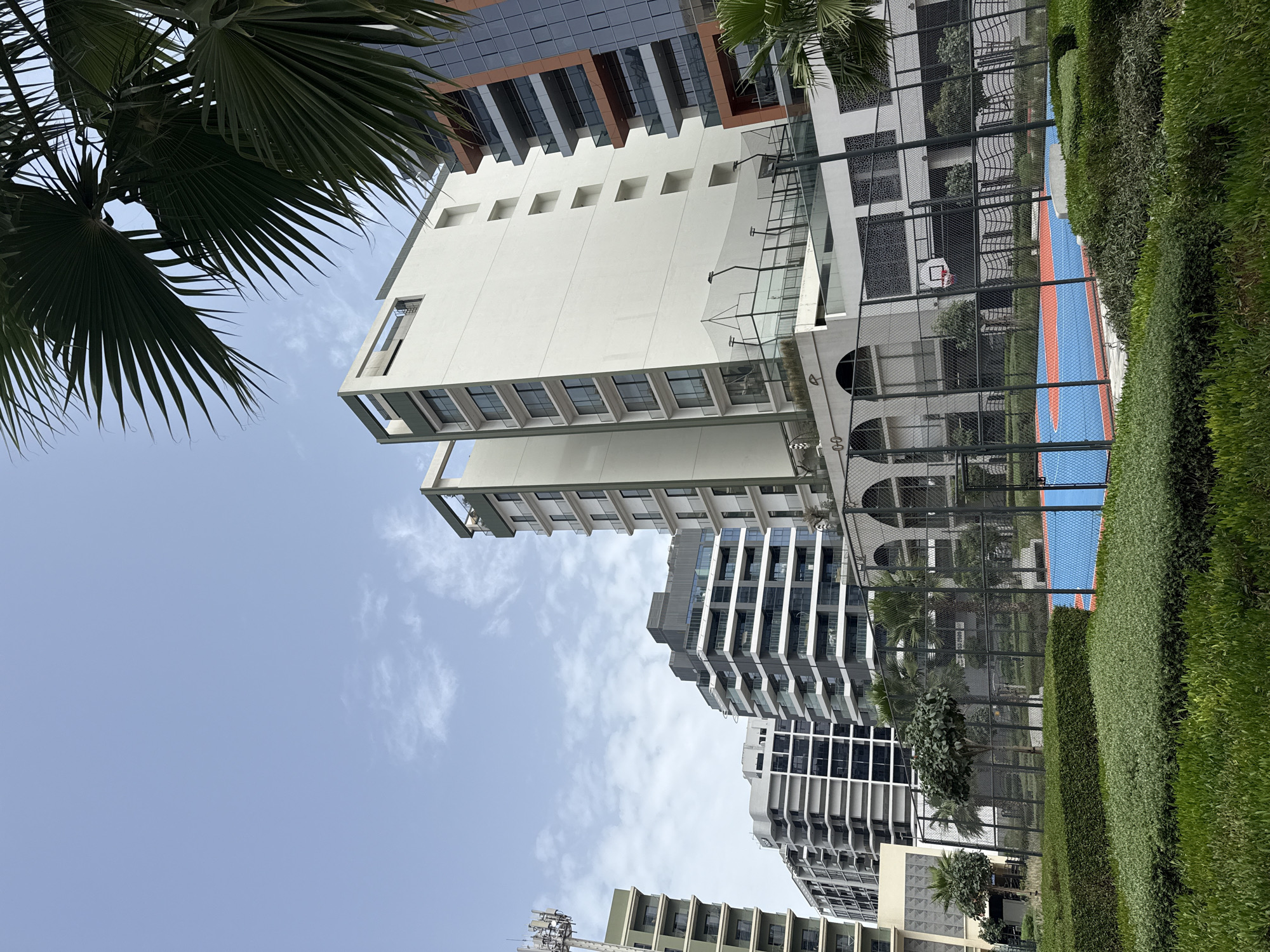 The image is a low-angle shot of buildings in Dubai, United Arab Emirates. The architecture is modern, with tall, multi-story buildings featuring geometric designs. A palm tree is visible in the foreground, partially obscuring the sky. A basketball court with blue and orange markings is surrounded by a fence and trimmed hedges. The sky is partly cloudy, with a blue hue. No people are visible. The image appears to be taken on a clear day.