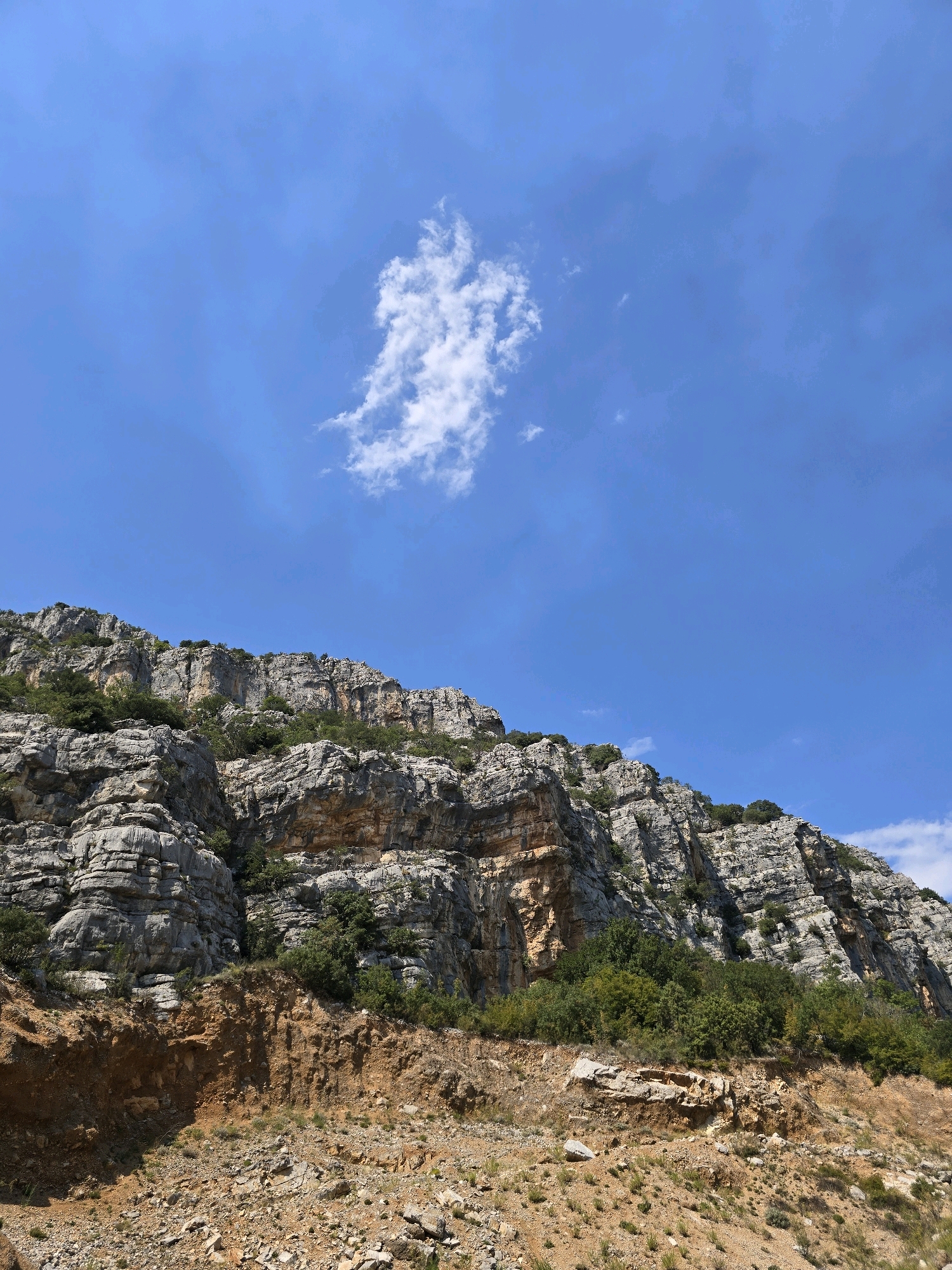 The image depicts a rugged, rocky mountainside under a clear blue sky with a few scattered white clouds. The rock formations are layered and appear weathered, with patches of green vegetation, including trees and shrubs, clinging to the slopes. In the foreground, a cutaway of the earth reveals exposed soil and loose rocks, suggesting a road or a construction site. The scene is devoid of any people or man-made structures, focusing solely on the natural landscape. While the specific location within Podgorica, Montenegro, is not explicitly identifiable from the image alone, it portrays a characteristic geological feature that could be found in the mountainous regions surrounding the city.