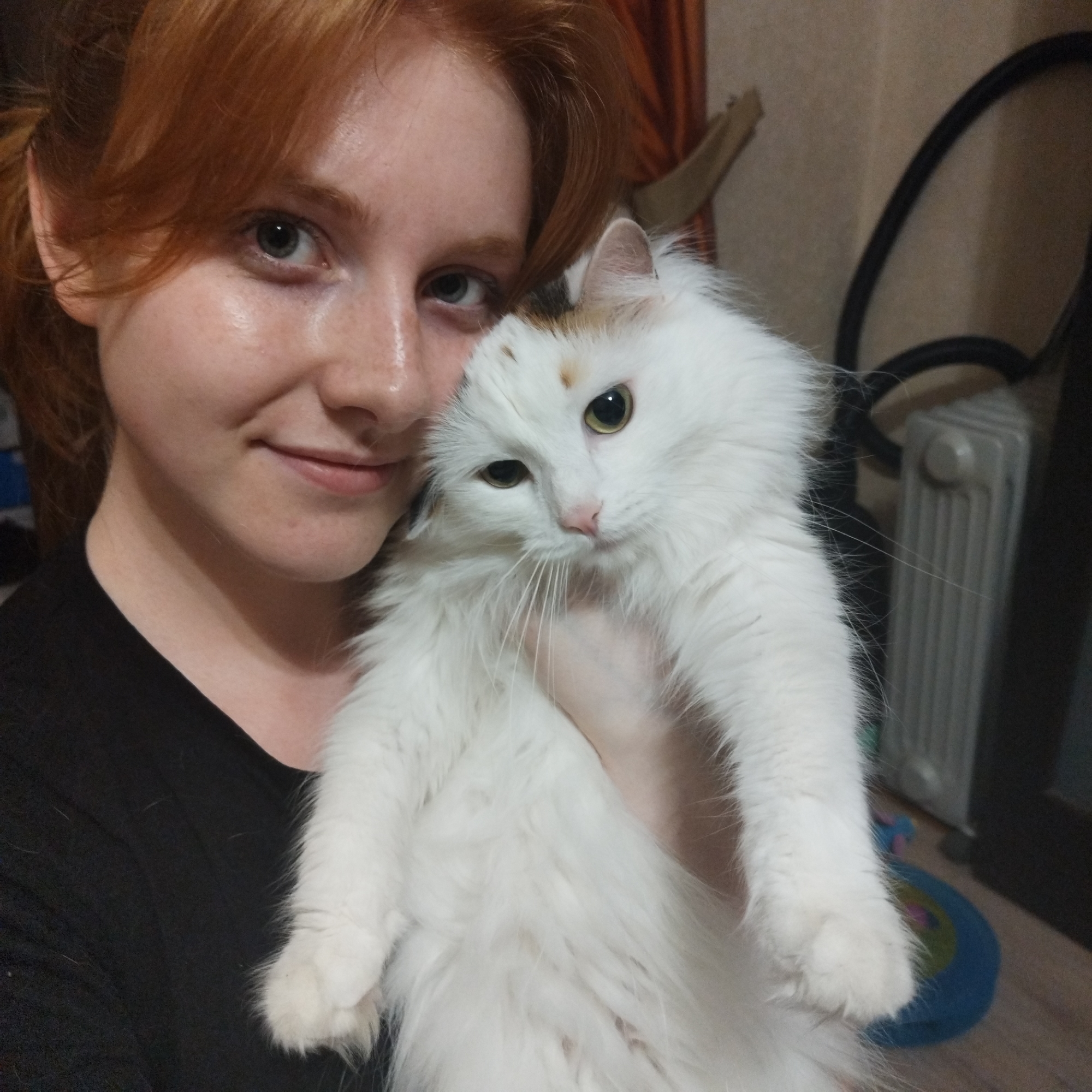A woman with red hair holds a fluffy white cat. The woman has fair skin and freckles and is wearing a black t-shirt. The cat has yellow eyes and a few spots of brown fur on its head. In the background, there is a radiator, a black cord, and a blue object on the floor. The location is likely indoors. No evidence suggesting the image was taken in Yekaterinburg, Russia.