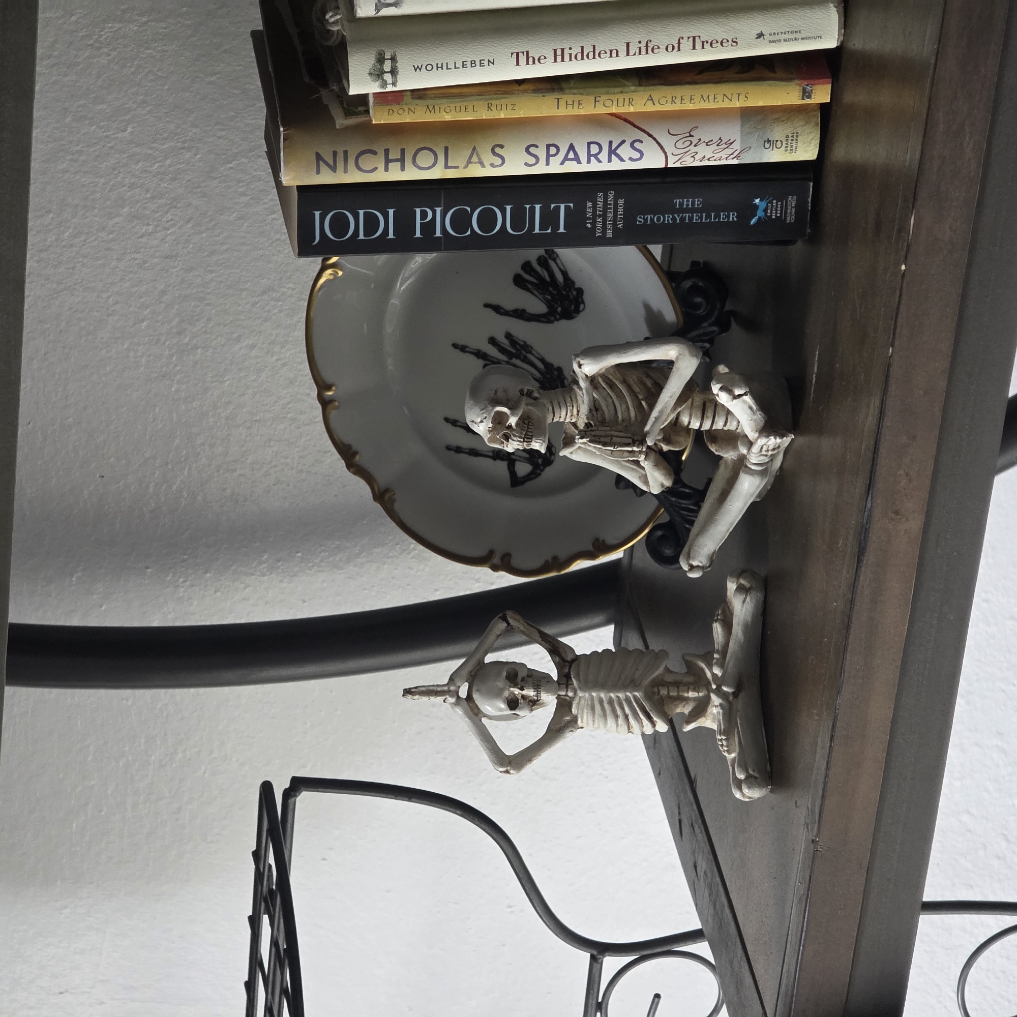 A shelf in an interior space displays decorative objects. On the left, a small skeleton figurine in a yoga pose is positioned on the shelf. To its right, another skeleton figurine is also in a meditative yoga pose. Behind the figurines, a decorative plate with a gold rim and black hand imprints is displayed. To the right of the plate, a row of books stands vertically. The shelf is made of dark wood. The wall behind the shelf is white. The background shows a dark, curved metal structure, possibly part of a shelving unit or furniture. The location is not specified in the image or its metadata.
