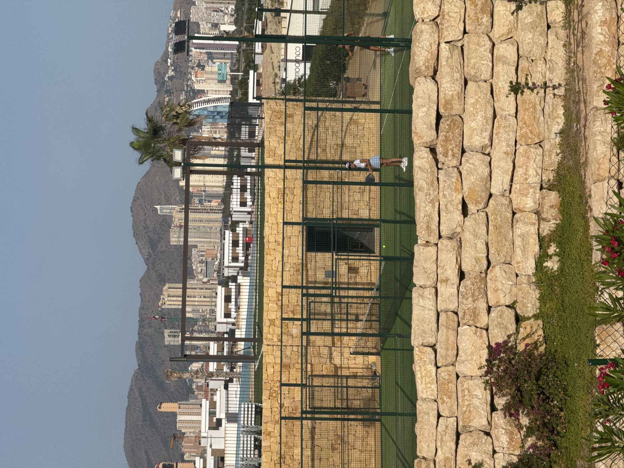 The image depicts a Padel tennis court in Finestrat, Spain. The court, with its green turf and glass walls, is situated in a scenic location overlooking a city and mountains. Two individuals are visible on the court, engaged in a game. One player, dressed in athletic wear, is positioned near the net, while another is seen further back on the court, potentially waiting for a shot or observing the game.
The surrounding environment suggests a sunny day, with a clear blue sky and bright lighting. The architecture of the city in the background, characterized by numerous buildings and structures, indicates a developed urban area. The presence of mountains behind the city adds a natural element to the landscape. A tall stone wall runs along the side of the court, a prominent feature of the setting. Green vegetation, including plants and flowers, grows along this wall, adding color and texture to the scene. The overall atmosphere appears to be one of recreation and leisure, set against a backdrop of striking natural and urban beauty. The word "coubocíaz" is visible on a sign in the background, though its context is unclear.