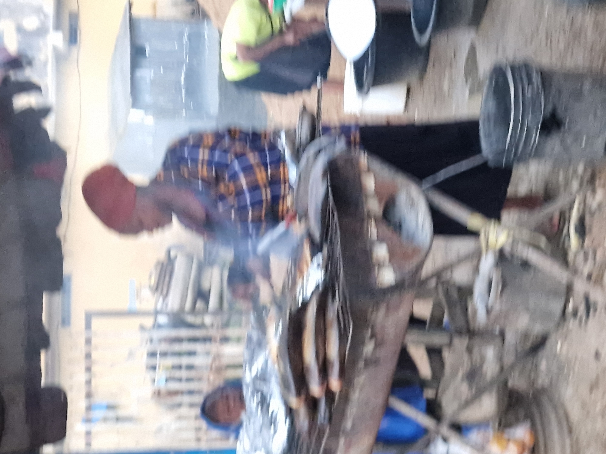 The image depicts an outdoor scene in Jos, Nigeria, likely a market or food stall area. A woman wearing a red head covering and a patterned blue and yellow blouse is actively grilling fish on a large, tiered metal grill. Several whole fish are visible on the grill, some covered in foil. Smoke emanates from the grill, indicating the cooking process is underway. In the background, a person wearing a blue head covering is partially visible, along with a light-colored building structure and a metal fence. Other elements in the scene include large black buckets and a white container. The ground appears to be unpaved. The overall atmosphere suggests a bustling commercial activity related to food preparation and sale.