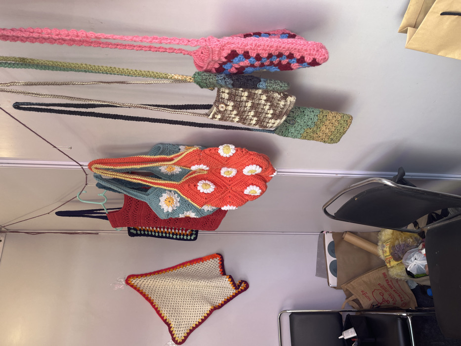 The image shows a variety of crocheted bags hanging on a string from a wall. The bags are all different colors and styles, and some have daisy-shaped designs. The bags are hanging on a wall in a room that contains a chair, a paper bag, and another crocheted piece. The location based on coordinates (latitude: 30.764, longitude: 76.798) is in Sector 17, Chandigarh, India.