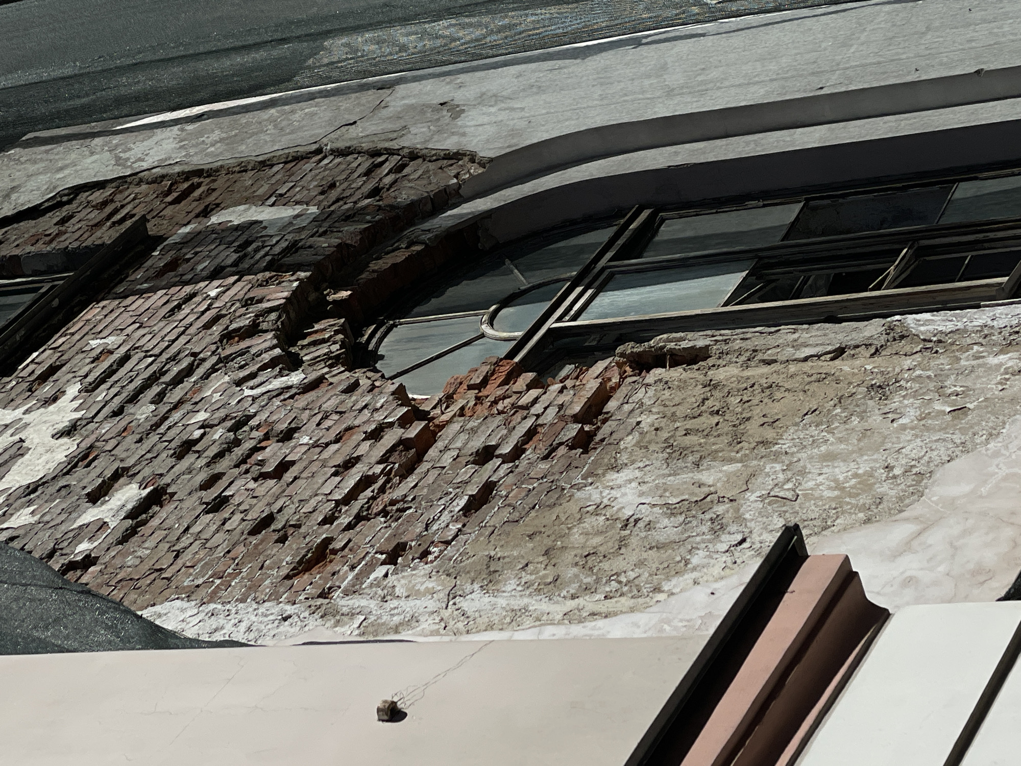 The image captures a severely dilapidated building facade in Saint Petersburg, Russia, seen from a slightly upward perspective. The primary subject is the building's weathered wall, where large sections of outer render or plaster have crumbled away, exposing irregular red and brown brickwork underneath. Patches of white residue or efflorescence are visible on the exposed surfaces, indicating long-term exposure and decay.
Several windows are visible on the facade. One prominent window features multiple panes, with some glass broken or missing, and a metal frame that appears bent outwards. Another window further along is partially open or damaged, showing a dark interior. The remaining plasterwork above the windows features a curved, decorative element, suggesting an older architectural style.
In the foreground, a light-colored, flat rooftop or ledge of an adjacent structure is visible, with a small, dark rock casting a shadow upon it. Above the damaged building, a dark, textured material, possibly a protective tarp or netting, is stretched across.
The scene is well-lit by direct sunlight, casting shadows and highlighting the rough textures of the brick and crumbling plaster, indicating daytime. There is no discernible human activity or visible text. The overall impression is one of neglect, significant structural decay, or damage over time in an urban setting.