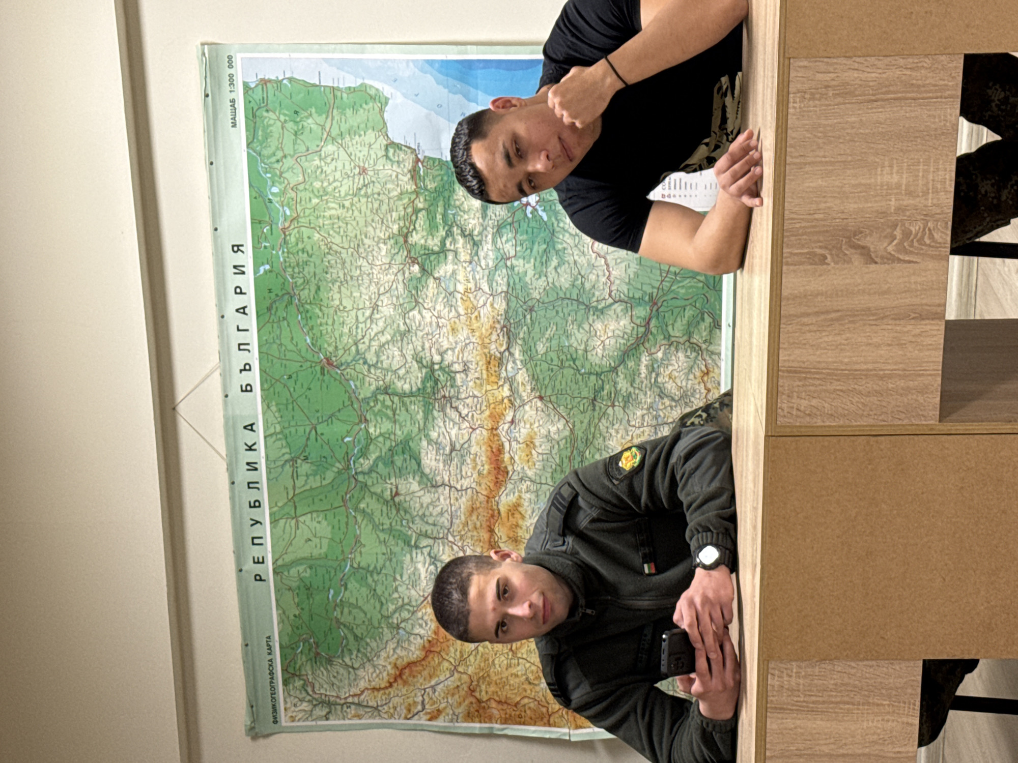 Two men are posing for a photograph in front of a large map of Bulgaria, with the phrase "РЕПУБЛИКА БЪЛГАРИЯ" (Republic of Bulgaria) visible on the map's left side. The map also includes text reading "ФИЗИКОГЕОГРАФСКА КАРТА" (Physical Geographic Map). One man is wearing a black t-shirt, and the other is wearing a dark green military-style jacket with patches and what appears to be a Bulgarian flag on the left sleeve. The man in the jacket is holding a phone in his hands. Both men are leaning on a desk or counter made of light-colored wood. The city and location are unknown.