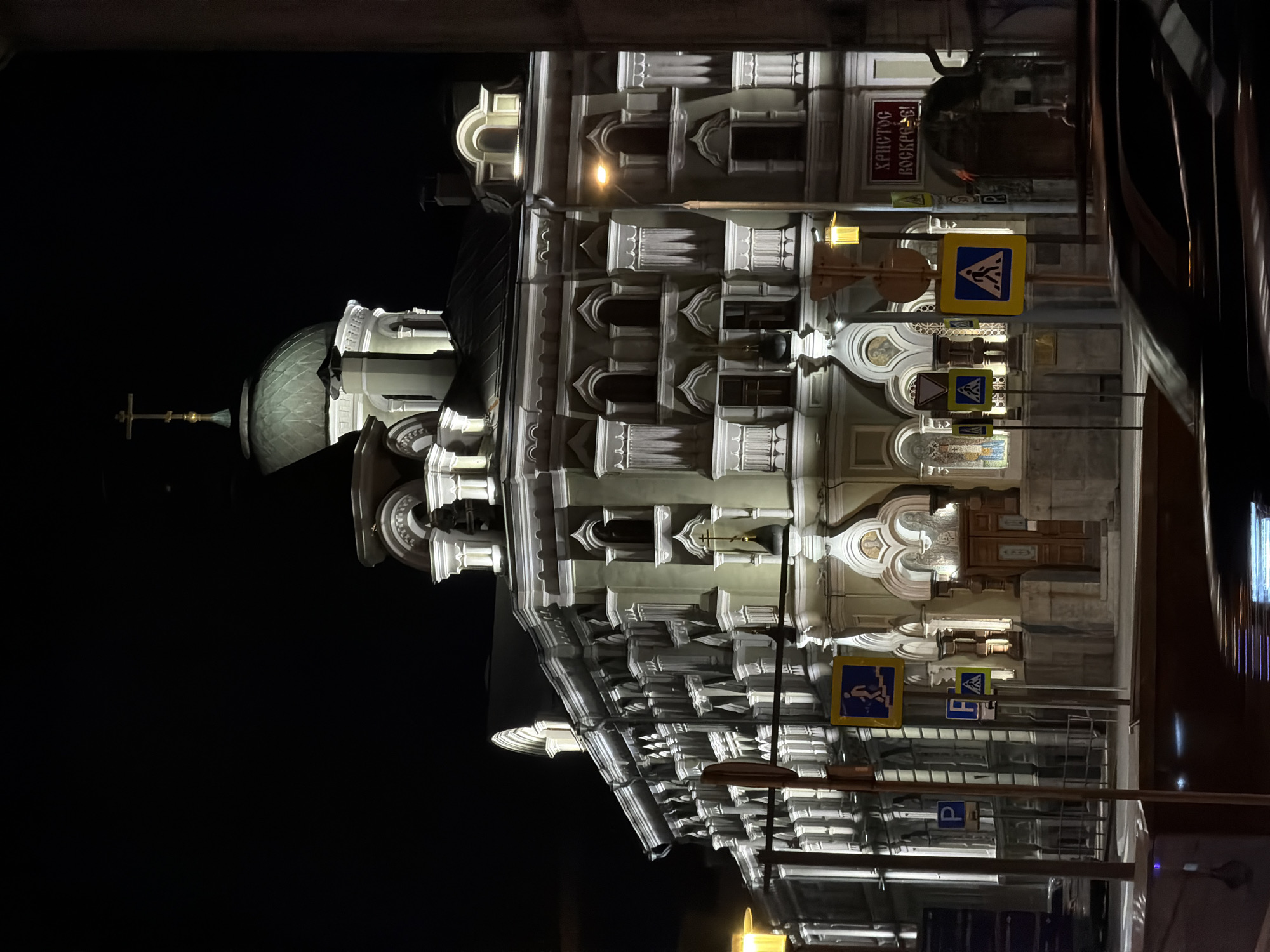 At night, a building is seen in Moscow, Russia. The building's facade is illuminated with white light, highlighting the architectural details, including decorative molding and arched windows. A dome with a cross tops the building. Several road signs, including pedestrian crossing and parking signs, are visible. The sky is dark.