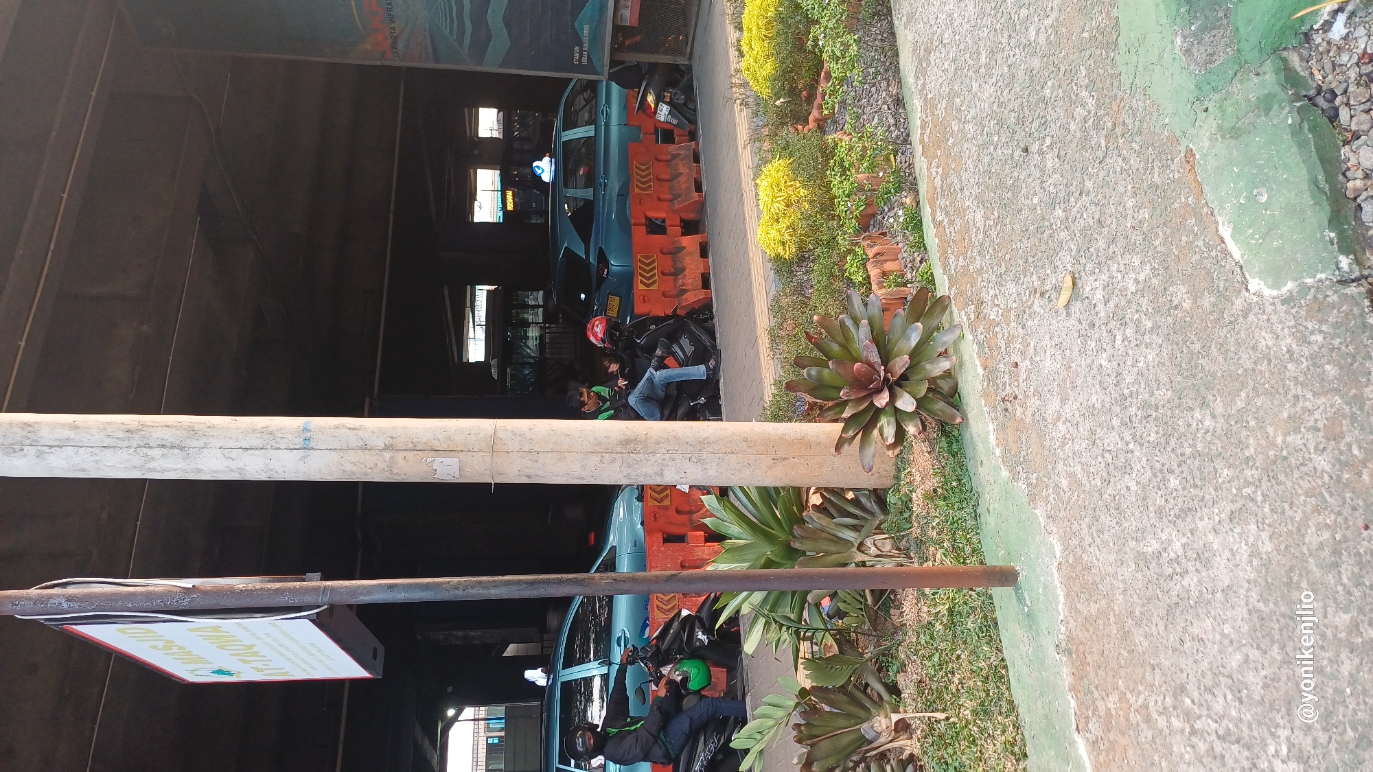 The image depicts an outdoor scene under an elevated structure, likely an overpass or parking garage, in South Jakarta, Indonesia. In the foreground, a concrete pathway is visible, bordered by a green painted curb and gravel. Several large plants with spiky, reddish-green leaves are growing in planters. A utility pole stands prominently in the mid-ground.
Behind the pole, two teal-colored cars with what appear to be taxi signs on their roofs are parked. Orange plastic barriers are placed in front of the cars, separating them from the planters. Two individuals are seated on black motorcycles parked behind the barriers and near the cars. The person on the left, wearing a black jacket and helmet, is on a motorcycle facing left. The person on the right, wearing jeans and a t-shirt, is on a motorcycle facing right, holding a phone. Another motorcycle is partially visible behind them.
A sign on a pole to the left of the frame reads "MASJID AT-TAQWA". In the background, the structure above creates a shaded environment, with some lighting and architectural elements visible. A poster or mural is partially visible on the right side of the image with text that includes "JAKARTA INFRA...". The overall impression is of a public space, possibly near a transportation hub or a mosque.