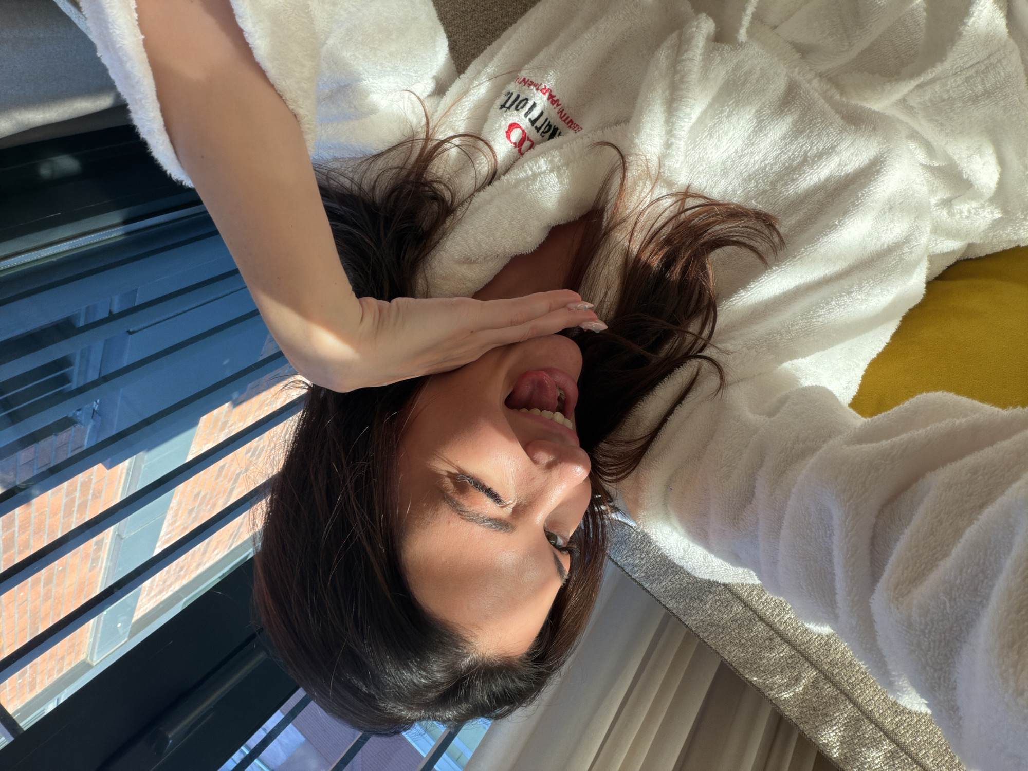 A woman with dark hair is lying on a light grey upholstered surface, possibly a bed or couch, wearing a white bathrobe with a "Marriott Apartments" logo in red and black. She is holding one hand up to her face, obscuring part of it, and sticking her tongue out. A window with metal bars is visible in the background, reflecting a brick building. Based on the logo on the bathrobe and the style of building visible through the window, it can be inferred that this photo was taken at a Marriott Apartments location, but it is impossible to state with any certainty if it is in the city of Küçükçekmece, Turkey.