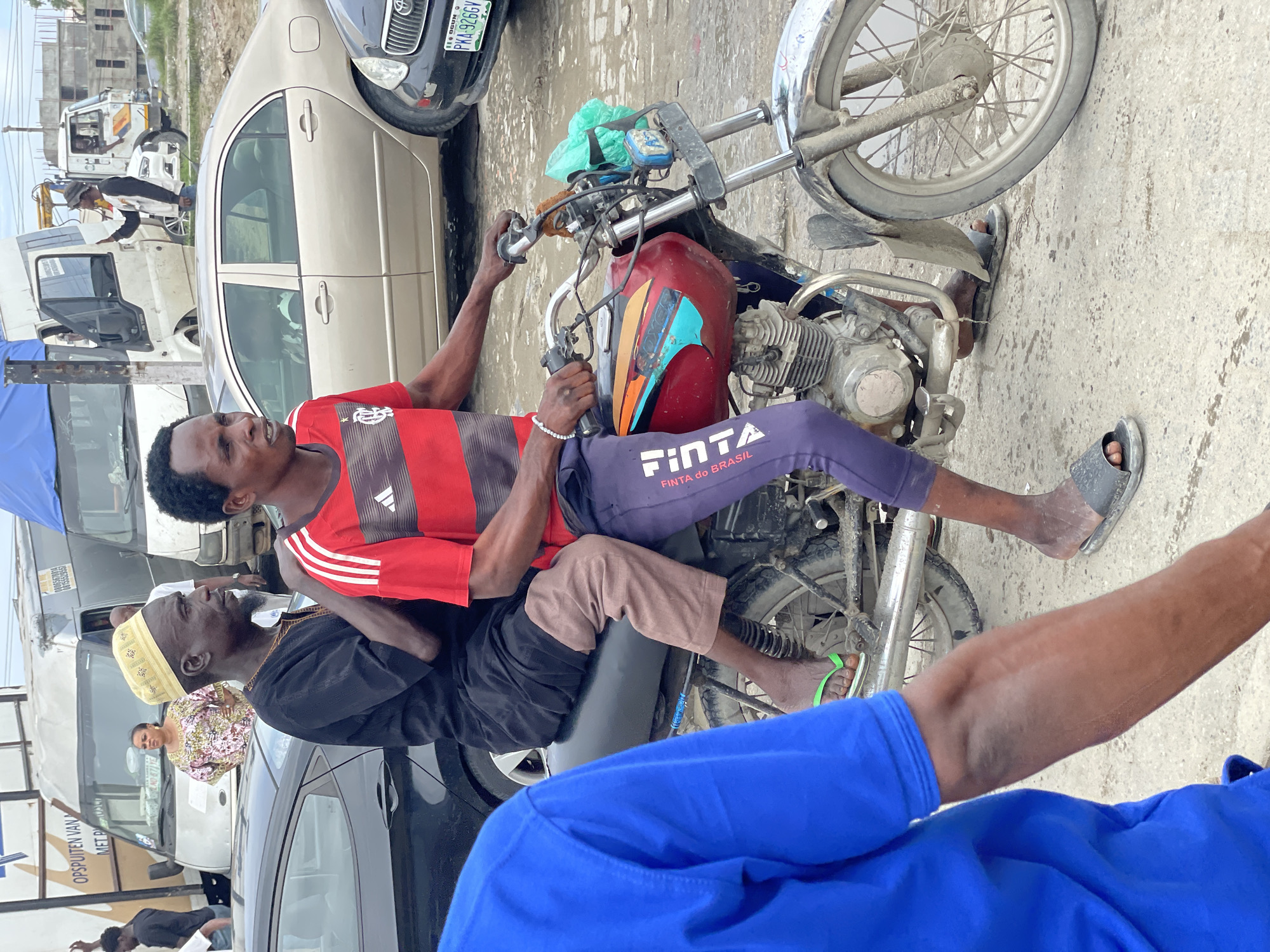 The image captures a street scene in Lekki, Nigeria, focusing on two men on a motorcycle. The man in the foreground, wearing a red and black striped t-shirt and dark pants, is looking to the left. He has a white beaded bracelet on his left wrist. The second man, seated behind him and facing right, wears a black robe and a yellow cap. He has a beard. The motorcycle is red with colorful graphics and appears to be in use, with a green plastic bag attached to the handlebars. The environment suggests an urban setting with paved roads, several vehicles including cars and vans, and some buildings or structures in the background. The ground is damp and appears to have puddles, indicative of recent rain or a wet environment. The overall impression is a candid moment of daily life in a busy Nigerian city.
