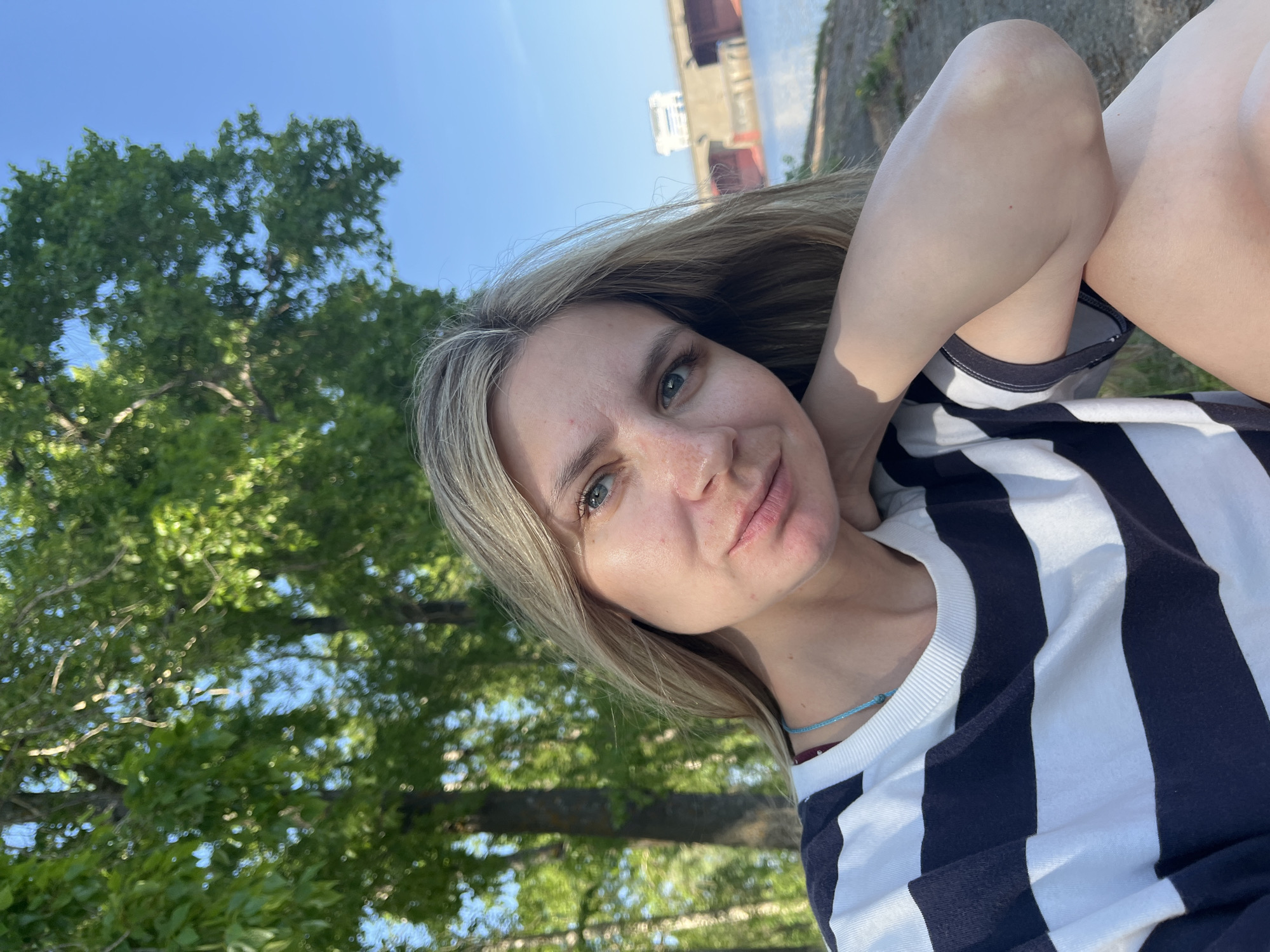 A selfie depicts a fair-skinned, blonde-haired woman in a striped shirt, likely taken outdoors on a sunny day in Chaikovsky, Russia. The woman is the primary subject, positioned in the foreground, with her arm raised behind her head. In the background, there are trees and blue sky. A building sits in the distance.