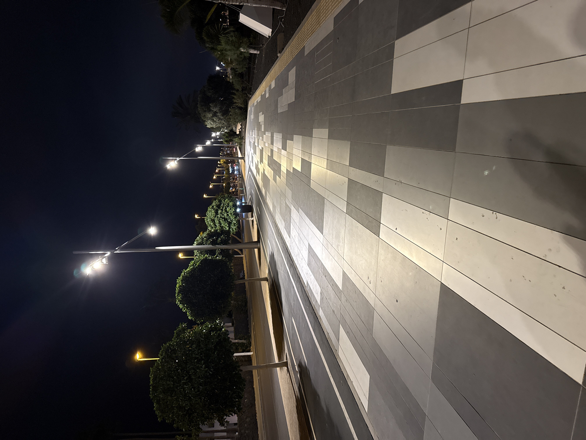 The image captures a nighttime scene on a pedestrian walkway in Tías, Spain. The walkway is constructed of alternating light gray and dark gray rectangular pavers, arranged in a pattern that creates a sense of depth. Streetlights illuminate the walkway and the adjacent area. On one side of the walkway, manicured trees are visible, and beyond them, additional streetlights and structures can be seen. The sky is dark, and the overall lighting is dim, with the streetlights providing the primary source of illumination.