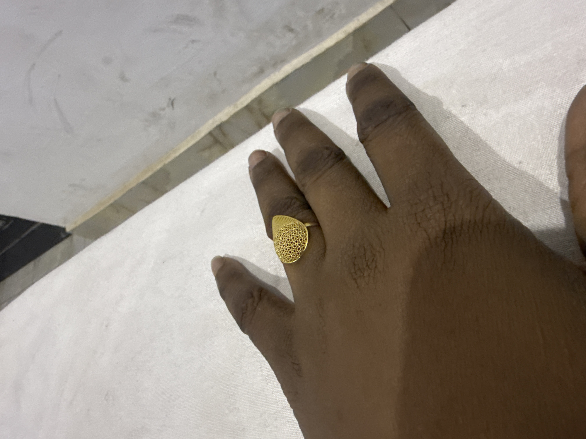 The image presents a close-up view of a person's hand, positioned on a white, textured fabric surface, likely a bedsheet or sofa covering. The hand exhibits a dark skin tone, with the fingers gently spread. A prominent gold-colored ring is worn on the ring finger. The ring features a distinctive, slightly elongated, teardrop or oval-shaped face adorned with an intricate filigree or perforated pattern, giving it a delicate, lace-like appearance.
In the background, behind the white fabric, a lighter, greyish surface is visible, possibly a wall or a piece of furniture, showing some minor smudges or imperfections. A thin, light-colored band, potentially the edge of a bed frame or furniture, separates the white fabric from this background surface. The lighting suggests an indoor setting, with some soft shadows cast by the hand. No discernible activity is taking place other than the hand resting. There are no other individuals visible, nor is there any visible text in the image. Based on the provided context, the image could be from Abuja, Nigeria.