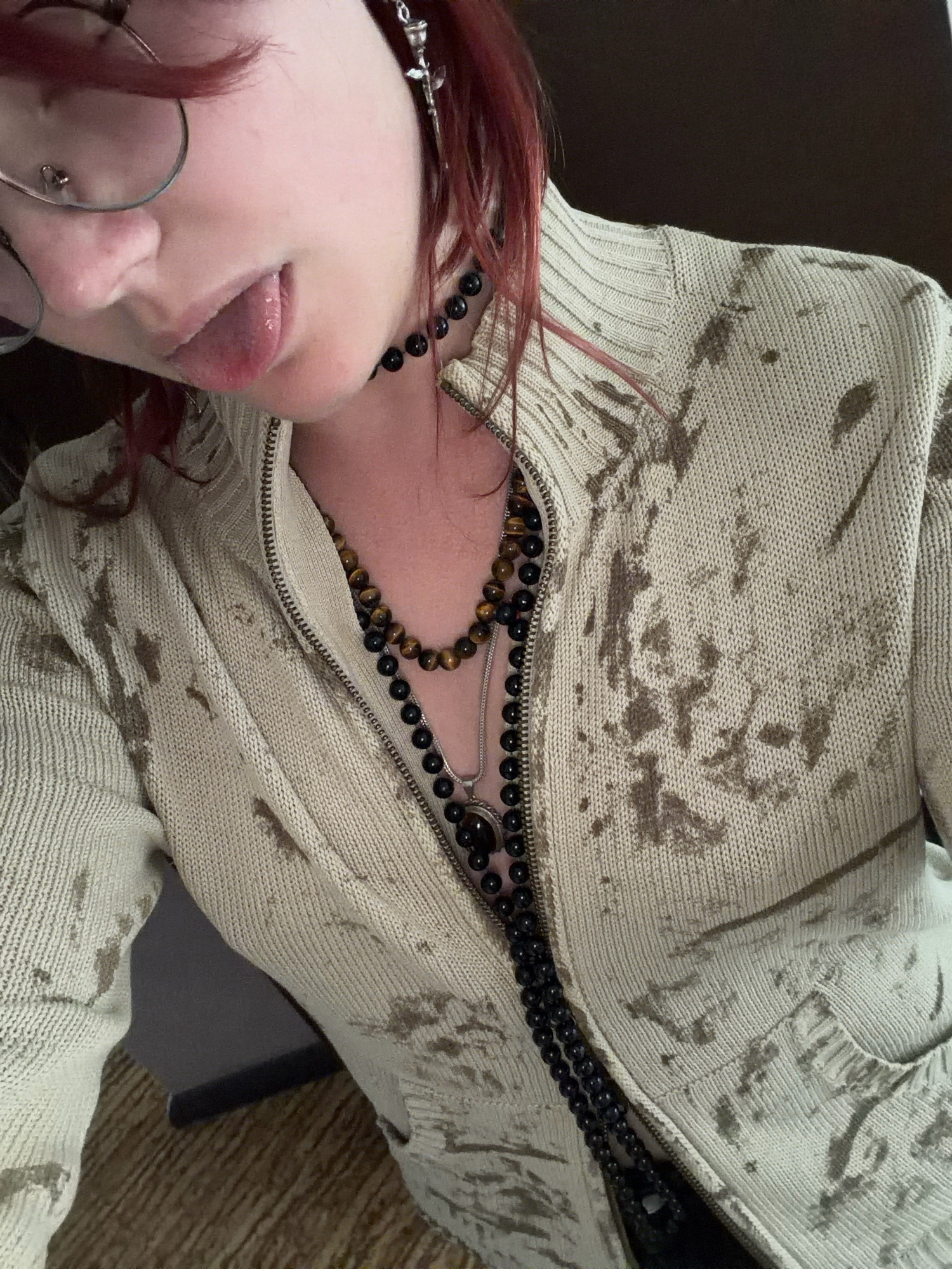 The image shows a close-up, medium shot of a person's upper body, viewed from a slightly high angle. The person has pale skin and reddish-brown hair. They are wearing clear-rimmed glasses with a nose ring, and their tongue is sticking out slightly, with the tip touching their lower lip. Around their neck, they are wearing two layered necklaces: a choker made of dark, round beads, and below it, a longer necklace with tiger's eye beads and a large, dark pendant.
The person is wearing a light-colored zip-up sweater with a textured, knit fabric. The sweater has a pattern of irregular, darker brown splotches across its surface. The zipper is partially undone, revealing the necklaces underneath. The background is dark and out of focus, with hints of a wooden surface at the bottom and a dark wall or fabric to the left.
There is no indication of the location being Chelyabinsk, Russia. The image appears to be taken indoors, possibly in a personal space. The focus is on the person's face, neck, and upper torso, emphasizing their accessories and the detail of the sweater.