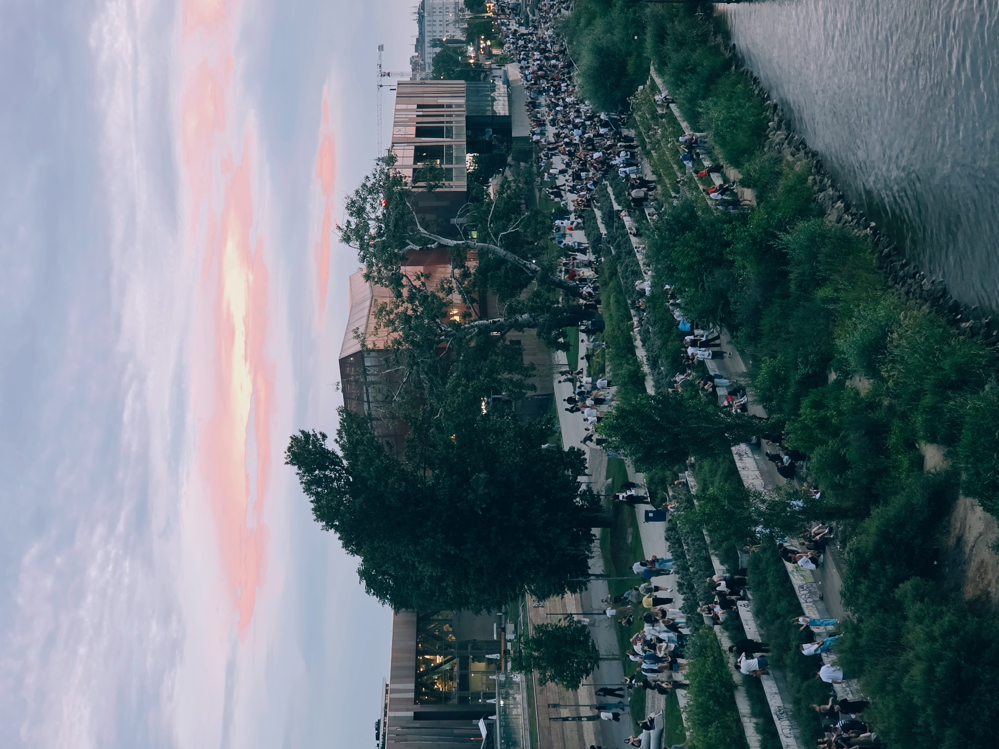 The image depicts a lively outdoor scene along the Vistula River in Warsaw, Poland, at dusk. A large crowd of people are gathered on tiered, landscaped seating areas that slope down towards the river. The people, dressed in casual attire, are engaged in various activities such as talking, sitting, and observing the surroundings. Many are seen walking along the pathways that wind through the terraced landscape.
Dominating the background is a modern, multi-story building with a distinctive copper-colored facade and large glass windows. The building appears to be a cultural or public facility, possibly a museum or entertainment venue. Adjacent to the building, a construction crane is visible, suggesting ongoing development in the area.
The sky above is a soft gradient of twilight colors, with streaks of pink and pale orange clouds against a muted blue backdrop. The river, a dark expanse of water, occupies the right side of the frame, with its surface reflecting the subdued light. Lush green vegetation, including trees and shrubs, lines the riverbank and the tiered seating areas, adding a natural element to the urban environment. The overall atmosphere is one of relaxed social gathering and enjoyment of a public space.
