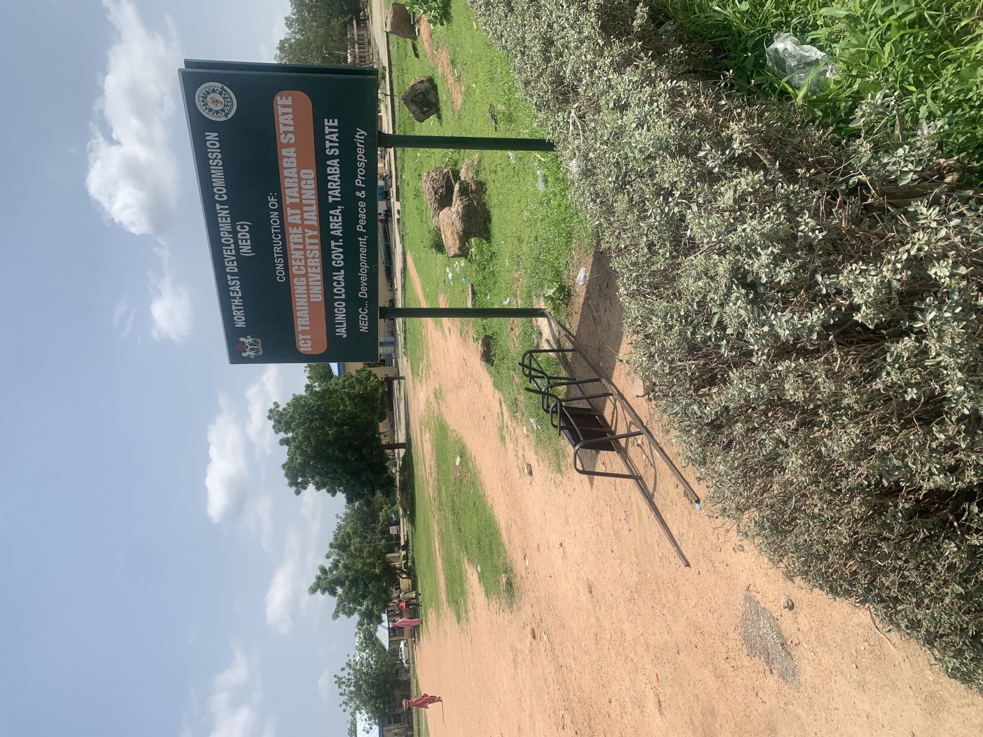 This is a medium-shot, outdoor, eye-level photograph of a construction site in Jalingo, Nigeria. The image is dominated by a large green sign in the center of the frame. The sign reads: "NORTH-EAST DEVELOPMENT COMMISSION (NEDC) CONSTRUCTION OF: ICT TRAINING CENTRE AT TARABA STATE UNIVERSITY JALINGO JALINGO LOCAL GOVT. AREA, TARABA STATE NEDC... Development, Peace & Prosperity." The sign is mounted on two metal poles, one of which is partially obscured by a dense green hedge in the foreground. To the left of the sign, a dirt path leads to an area with sparse grass and a few trees. In the background, several buildings with blue roofs are visible, along with a few people walking. One person, seen from the side, wears a pink garment and carries a basket on their head. Another person in a light-colored outfit walks away from the camera. The overall environment appears to be a semi-arid region, with dry, sandy soil and scattered vegetation. A metal structure, possibly a bicycle rack, is visible in the foreground, to the left of the sign. The sky is overcast with scattered clouds.